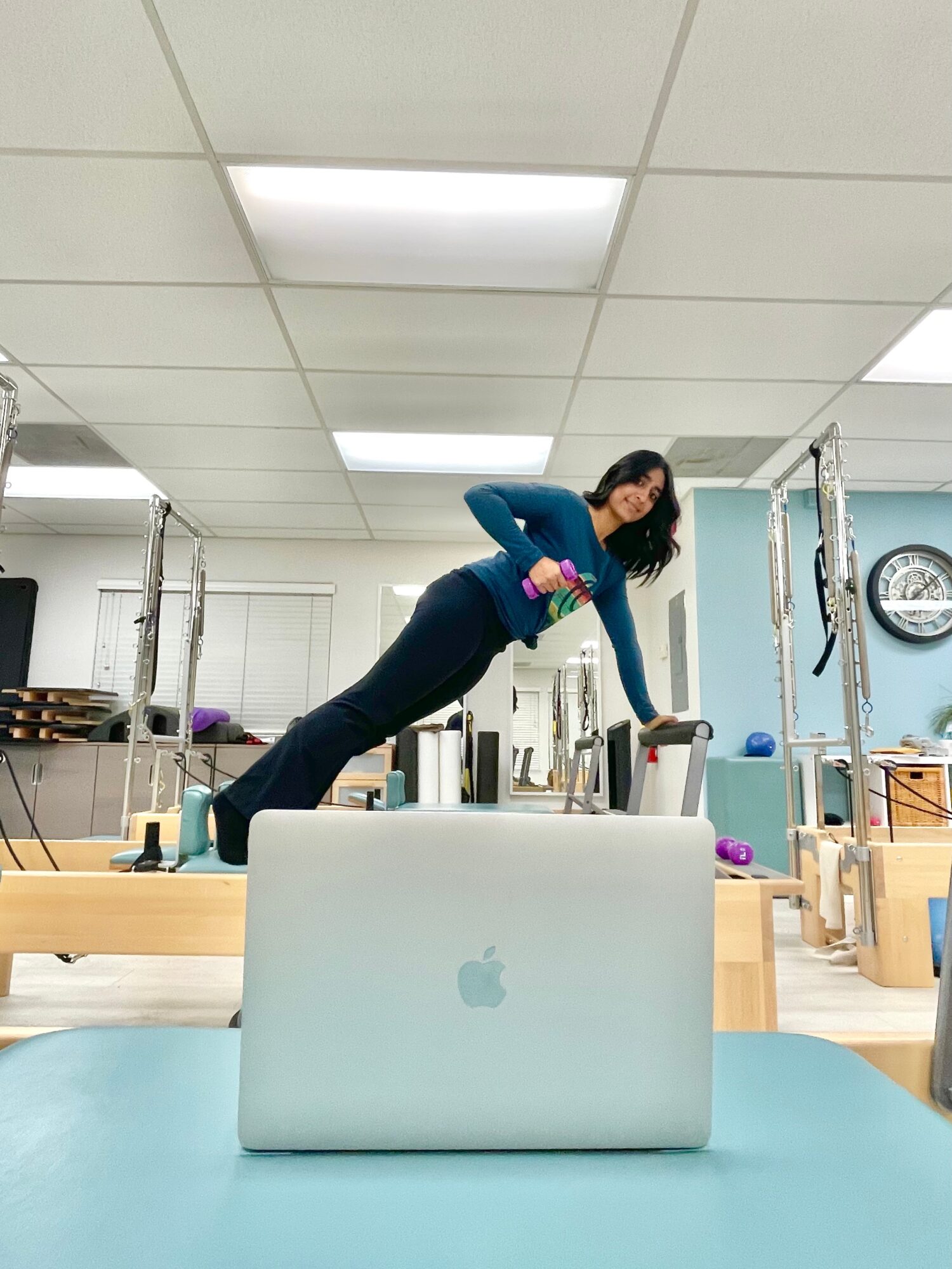 Woman in workout clothes performs a side plank in a fitness studio, with a laptop in the foreground.