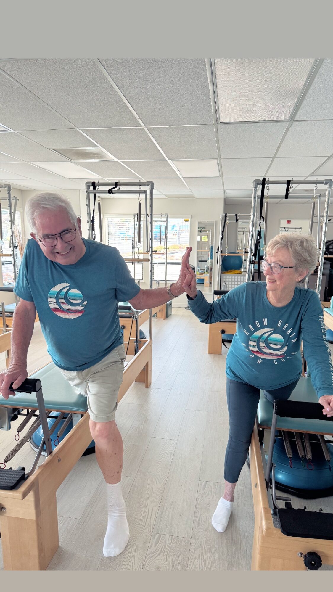 Two elderly people in matching blue shirts perform a high-five in a rehabilitation center, with exercise equipment around them.