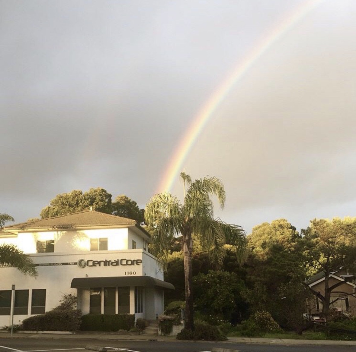 Rainbow arching over a building and trees on a cloudy day, with sunlight illuminating the scene.