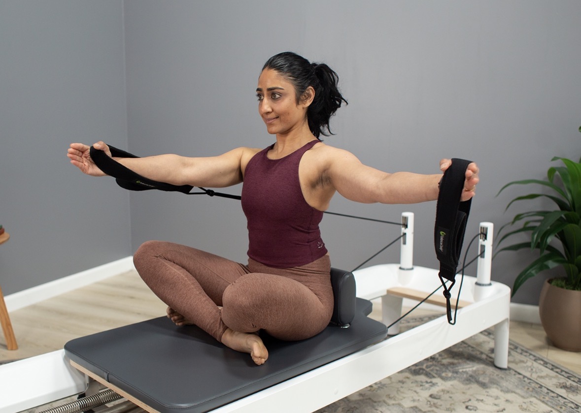 Woman sitting cross-legged on a platform, using resistance bands for exercise, in a room with gray walls and a plant.