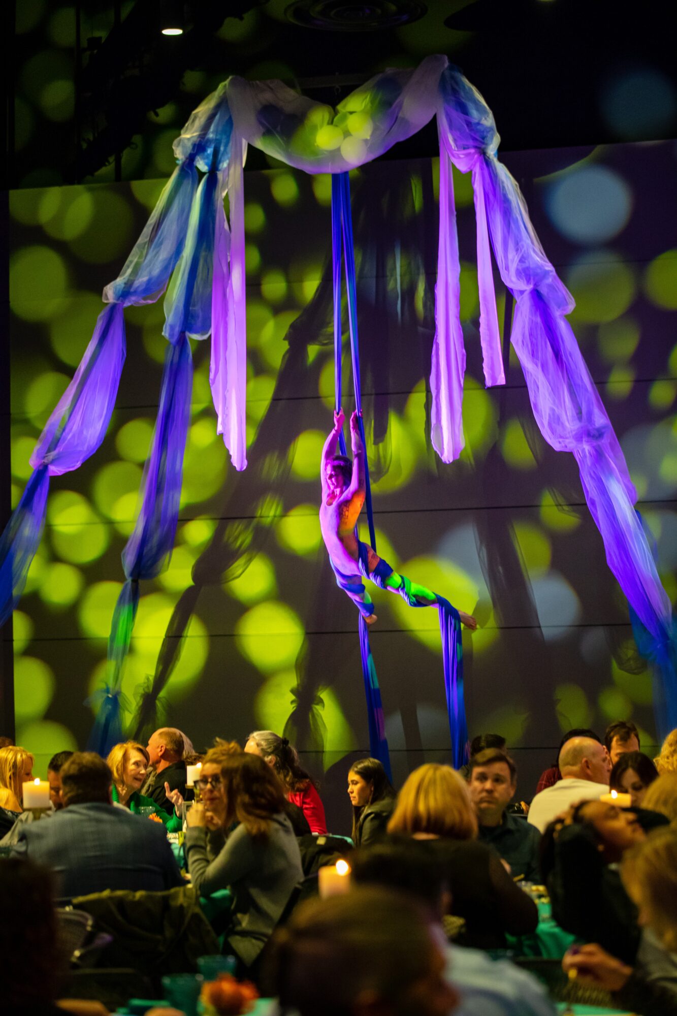 Colorful hanging fabric art installation above a seated audience in a dimly lit room.