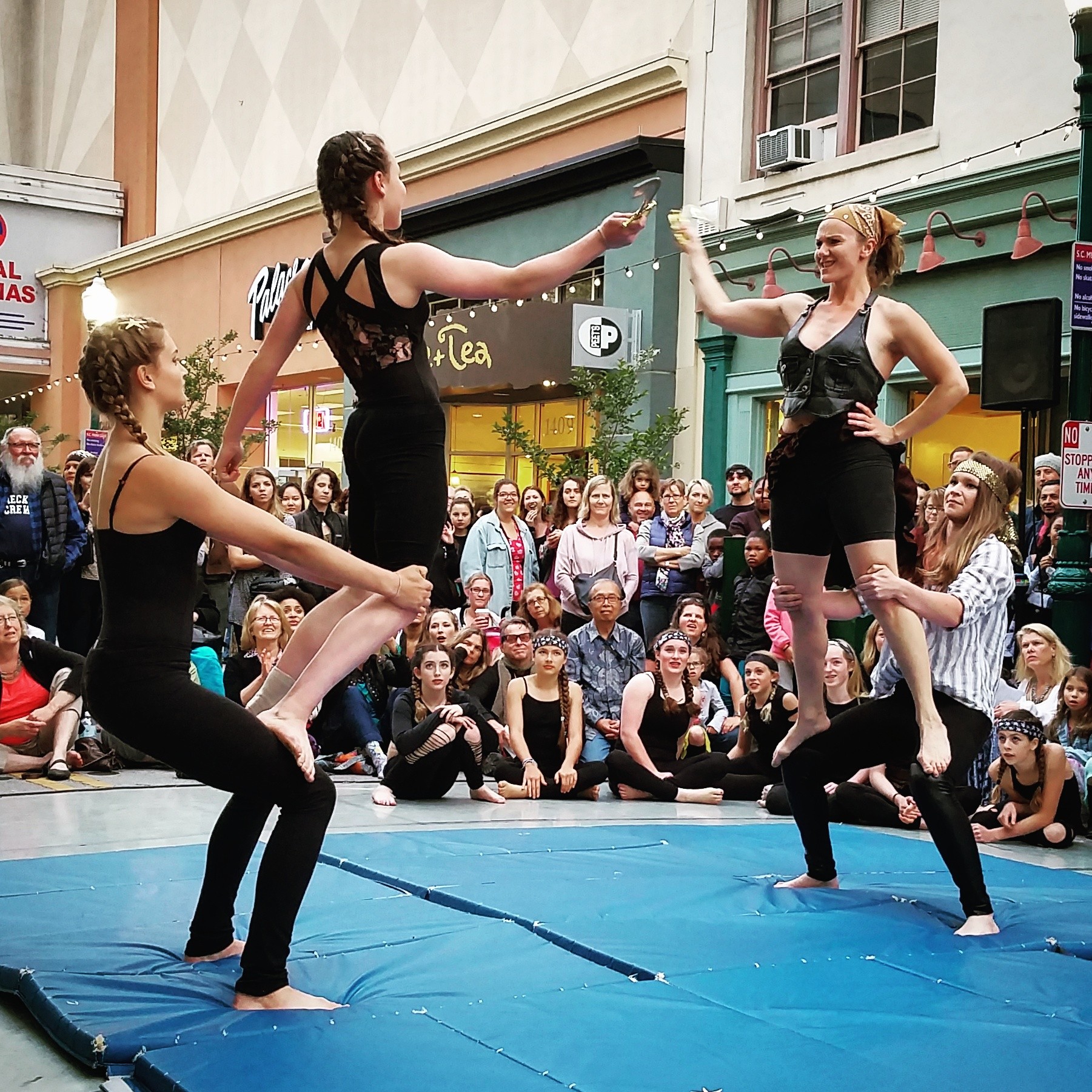 Three performers in black costumes perform a balancing act on a blue mat, with a crowd watching outdoors.