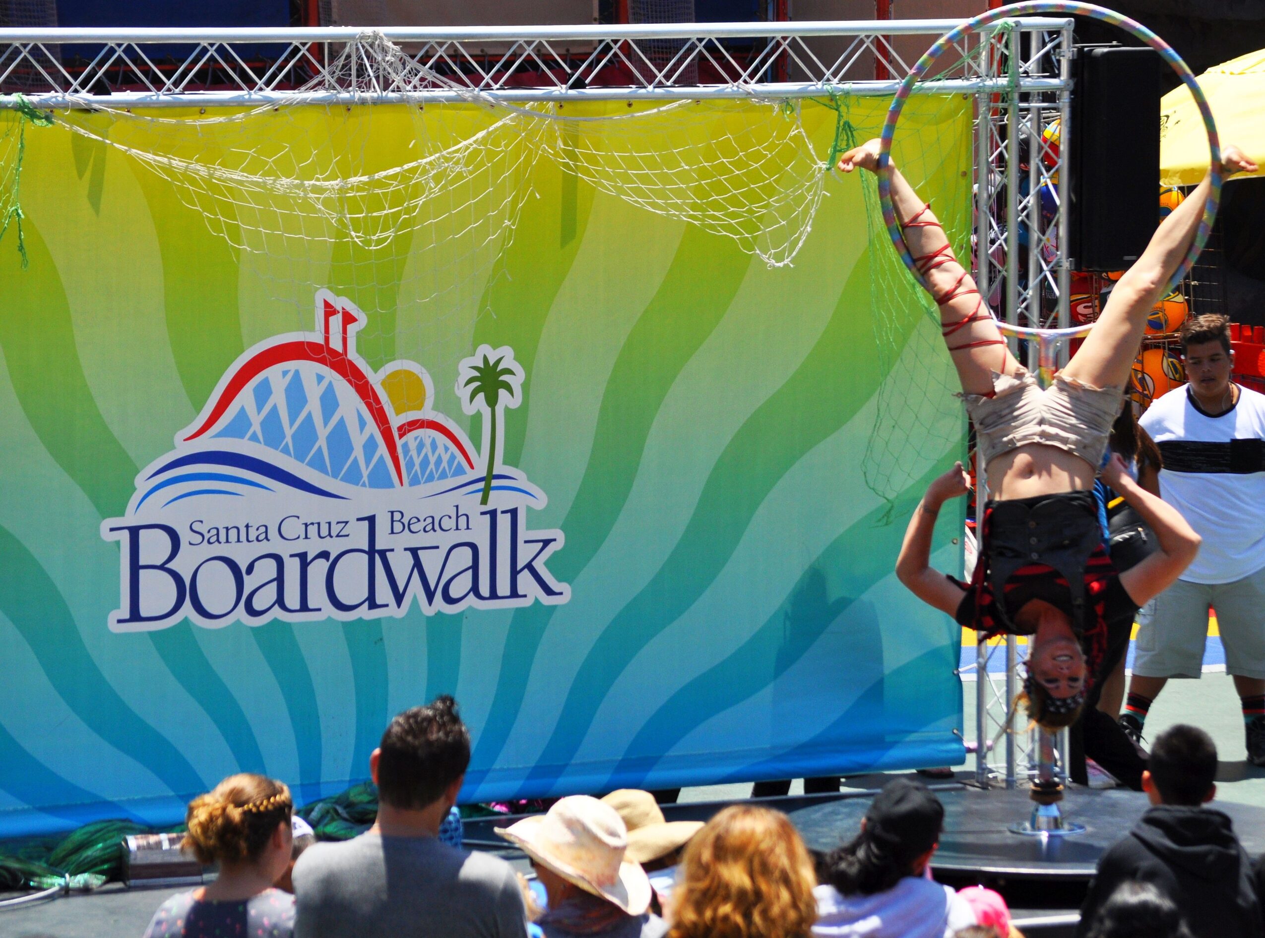 A performer hangs upside down on a hoop during a circus act on stage at Santa Cruz Beach Boardwalk, with an audience watching.