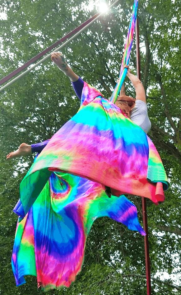 Person on aerial silk in colorful tie-dye fabric, trees and sunlight in background.