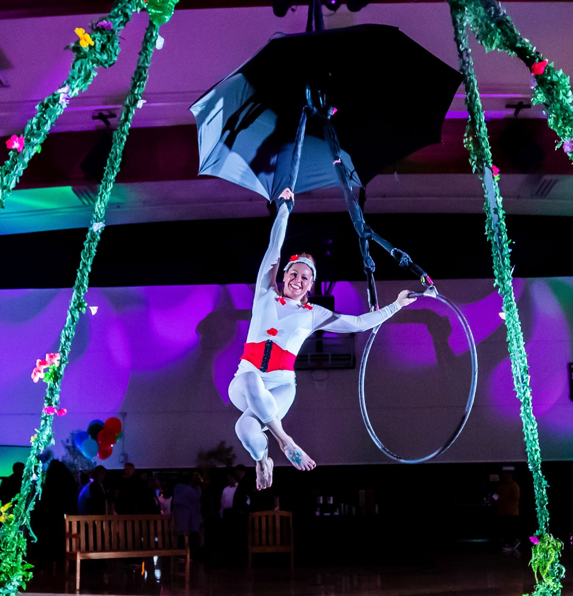 Circus performer hanging from a trapeze with a black umbrella, surrounded by colorful decorations, in a decorated indoor venue.