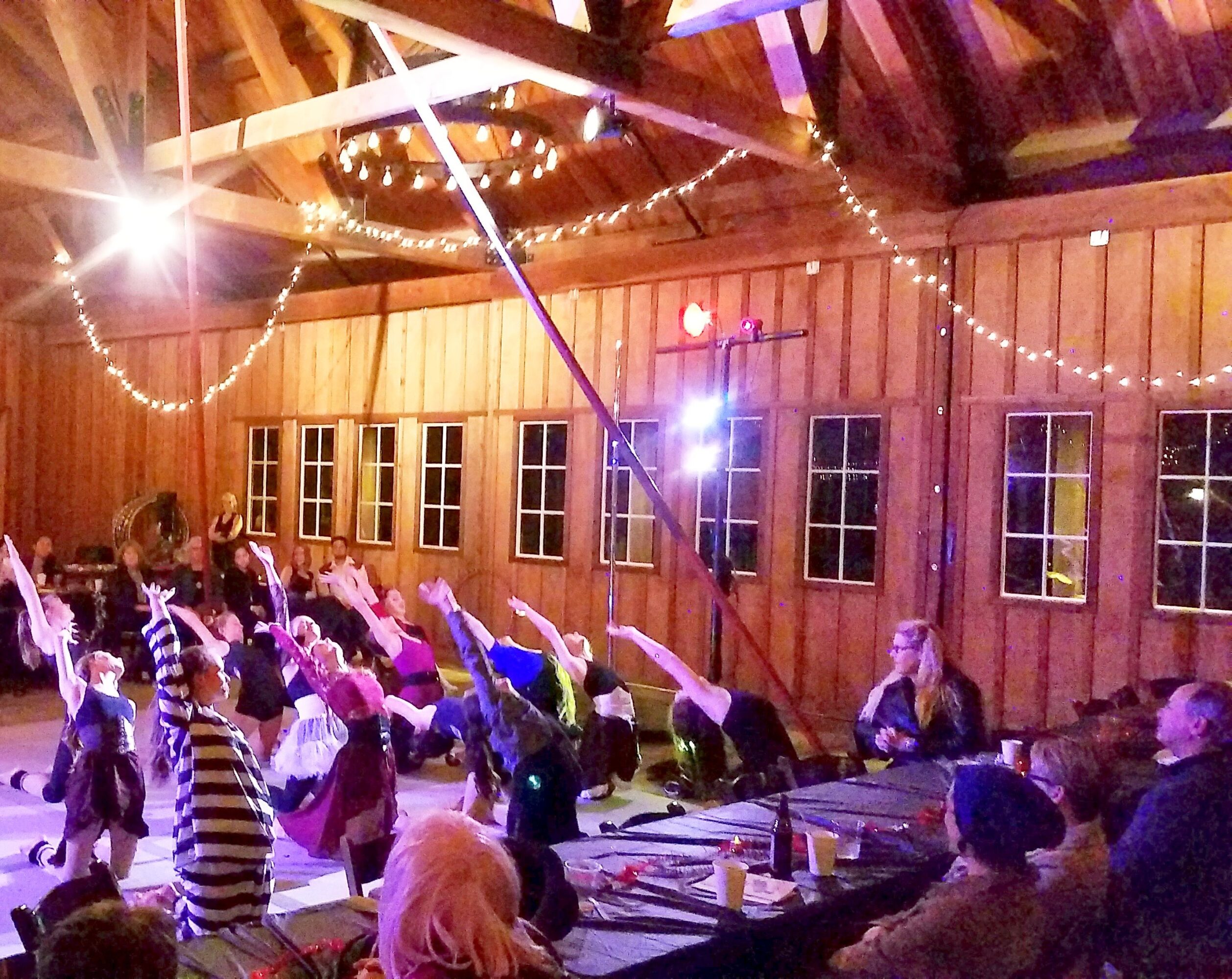 Children perform a dance with arms raised in a wooden hall decorated with string lights and a circular light fixture.