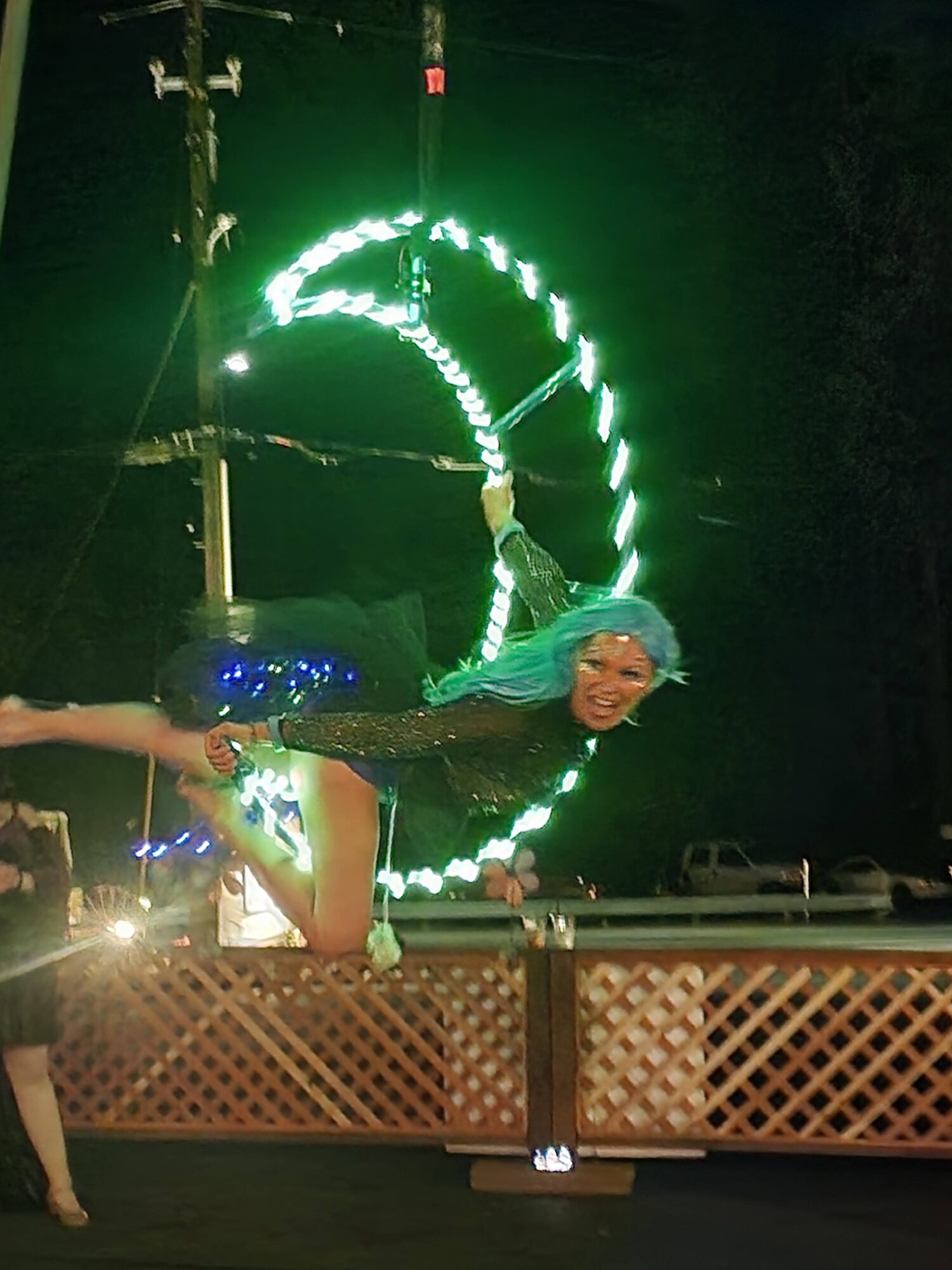 Performer in costume with blue hair and bright outfit, balancing on a railing at night with a lit Ferris wheel in background.