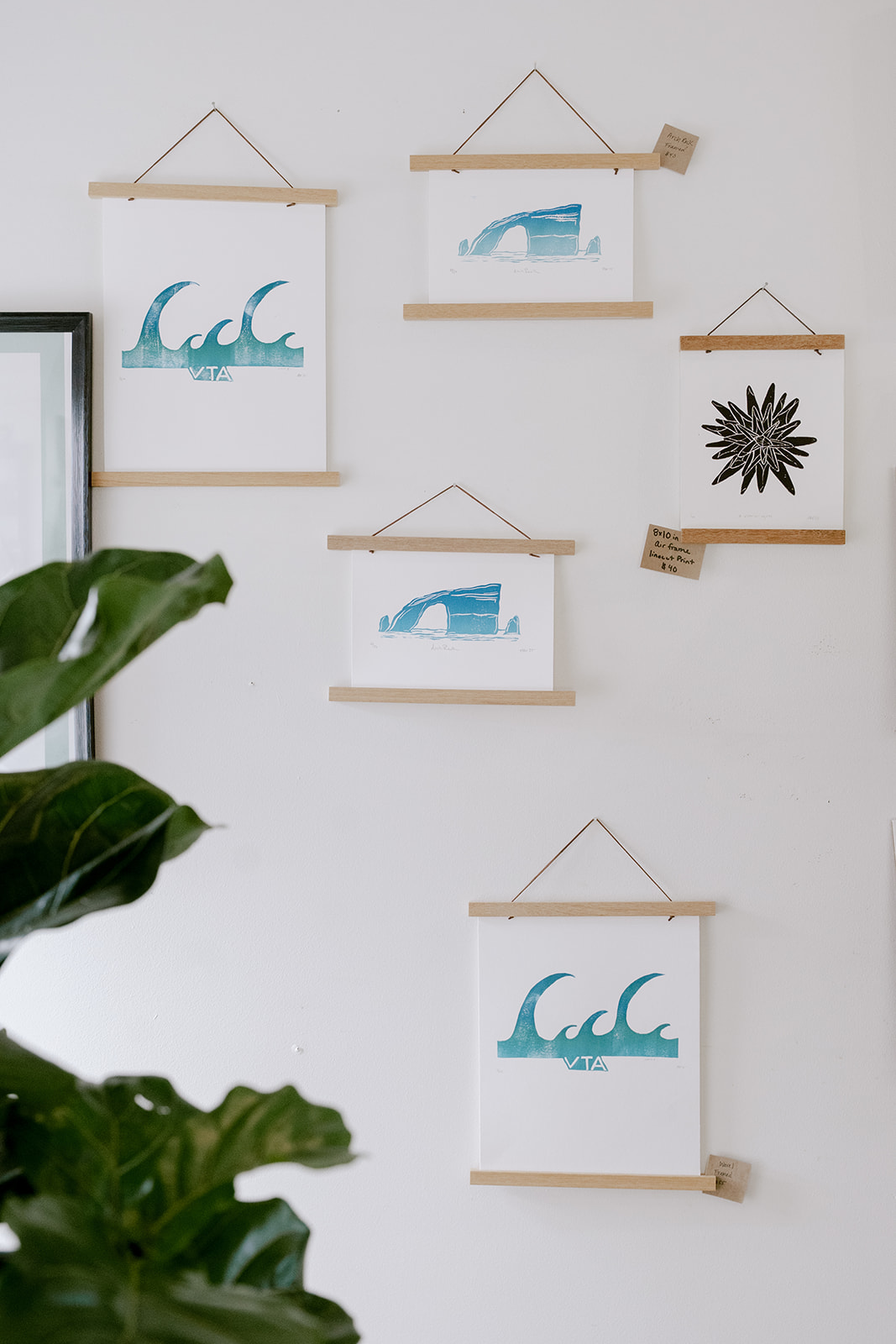Wall with five framed art pieces, some with blue wave designs, and a black decorative element, partially obscured by a green plant.