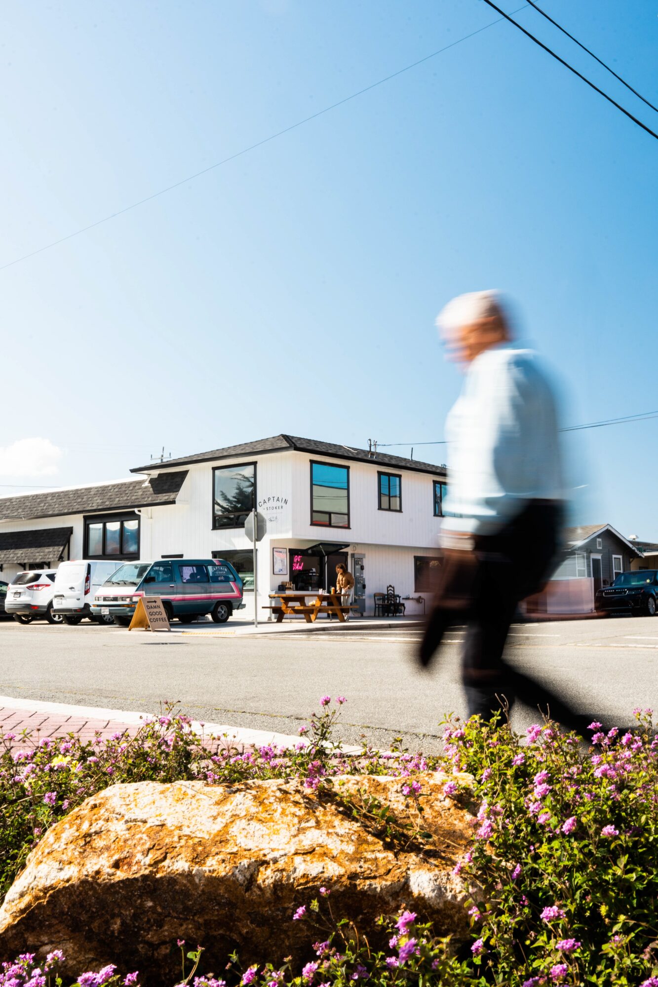 Person walking past a building with parked cars, flowers, and rocks in foreground, under a clear blue sky.