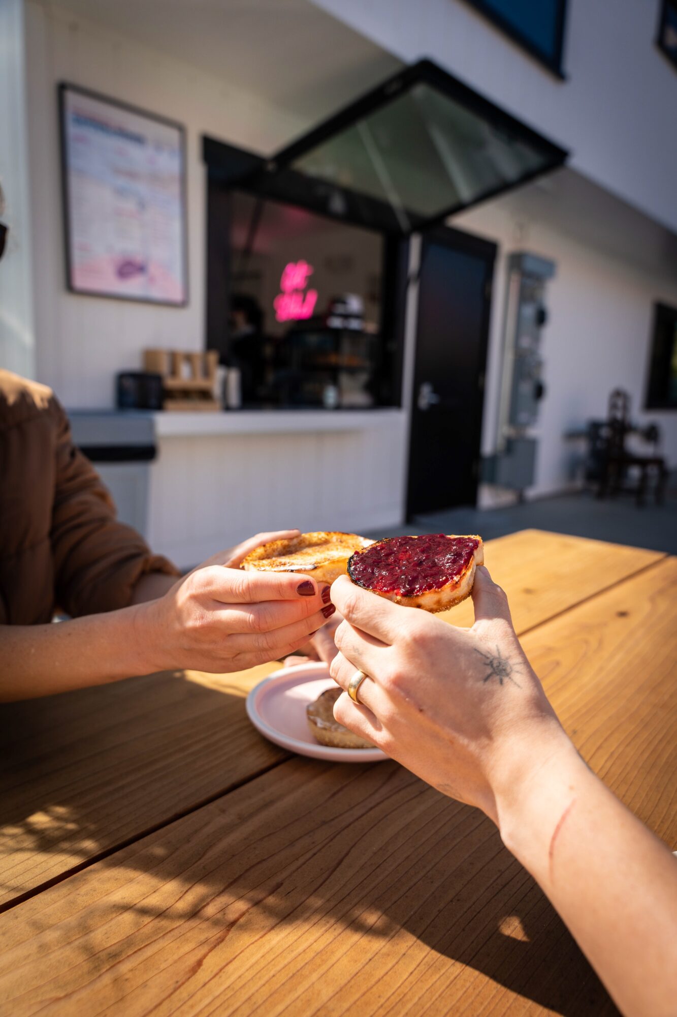 Two people exchange slices of cake at a wooden table inside a cafe, with a counter and menu in the background.