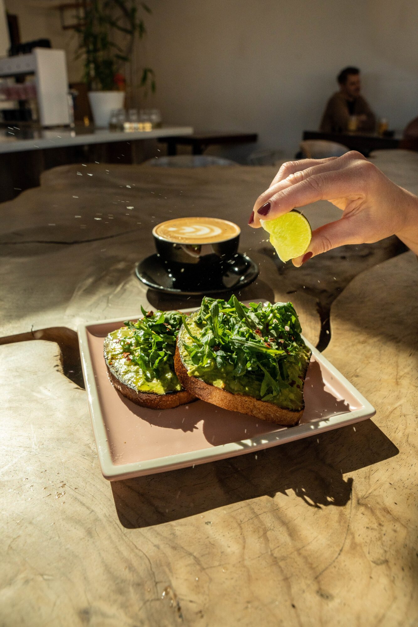Hand squeezing lemon over avocado toast on a pink plate, with a cup of coffee in the background.