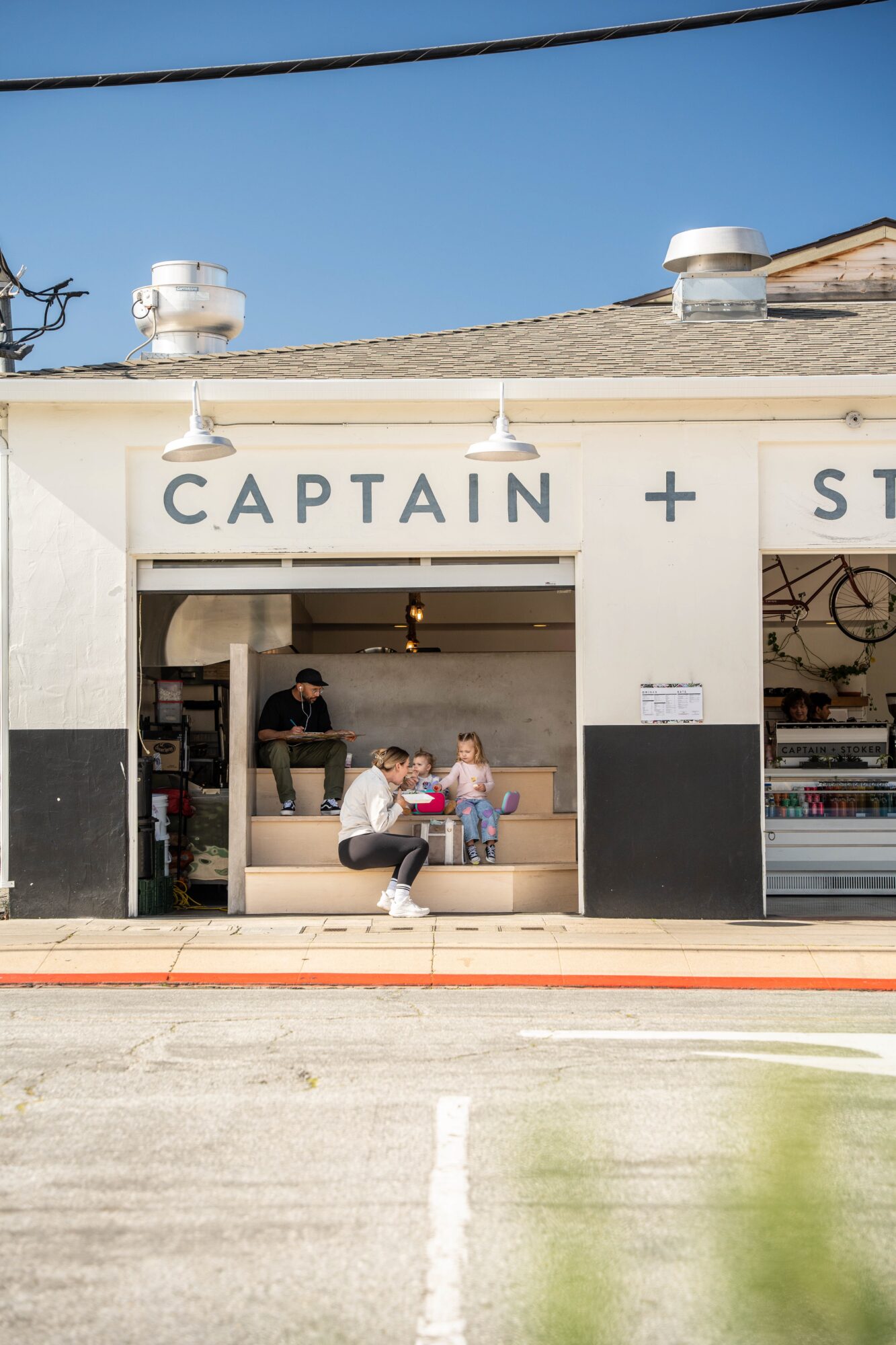 People sitting outside a storefront with a sign reading 'CAPTAIN + S', under a clear blue sky.