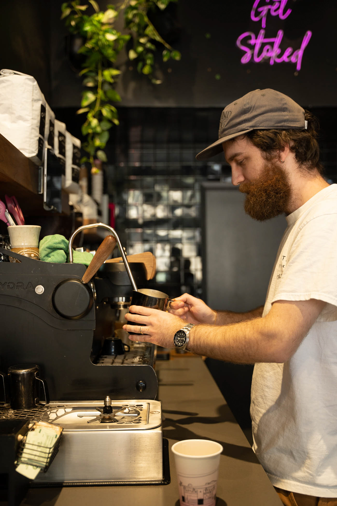 Barista preparing coffee behind the counter with a neon sign reading 'Get Stoked' in the background.