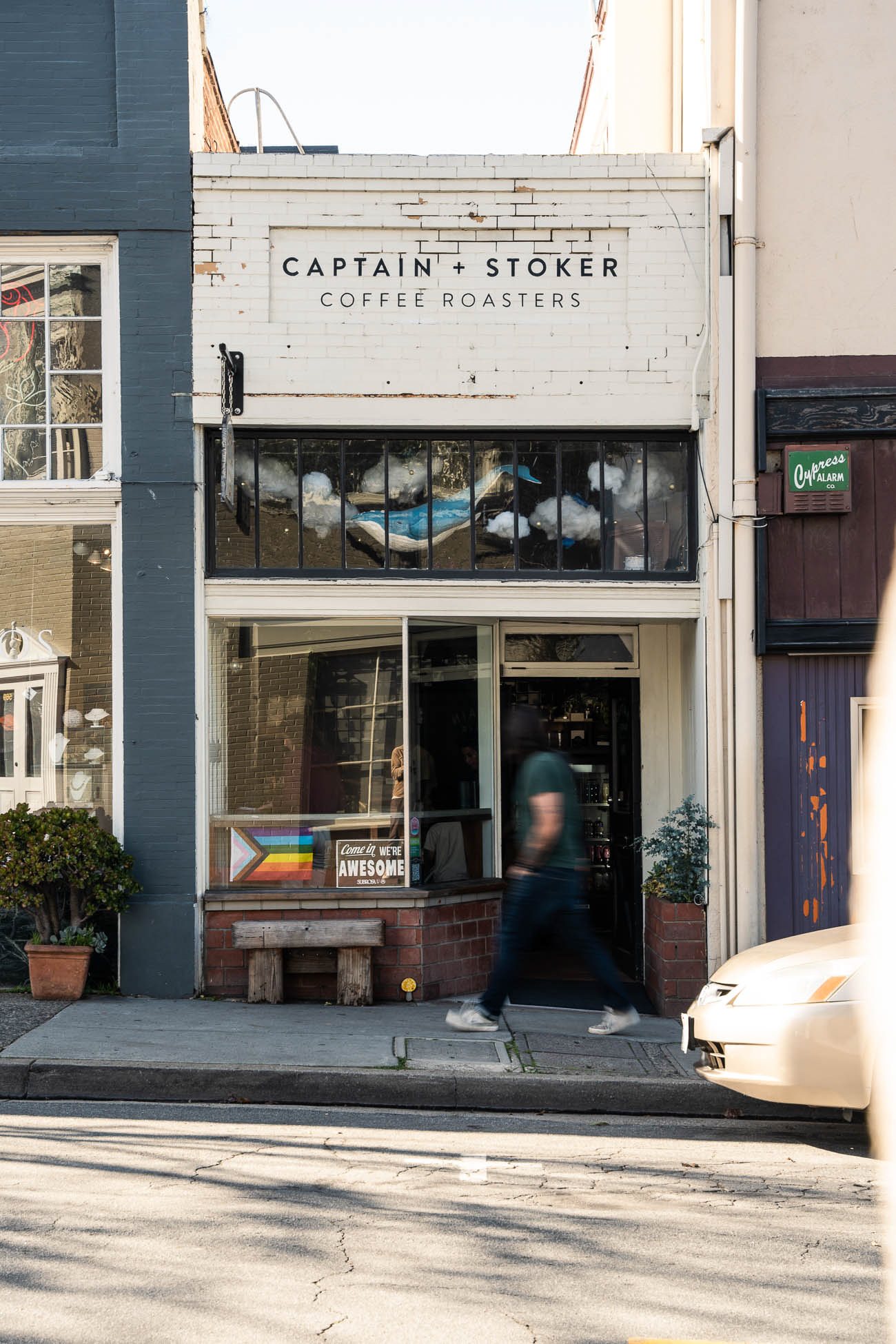 Street view of a coffee shop with a sign reading 'Captain + Stoker Coffee Roasters,' a window, and a person walking by.