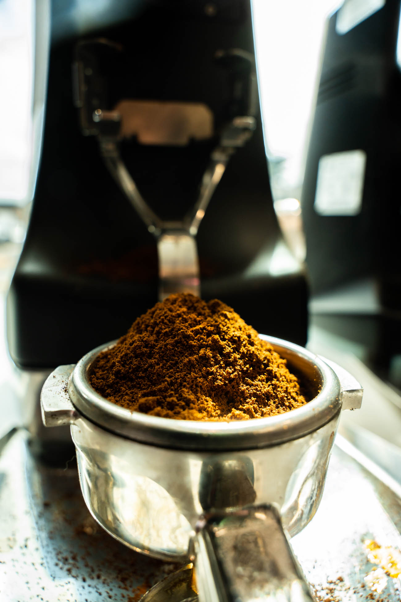 A portafilter filled with ground coffee, with a coffee machine in the background.