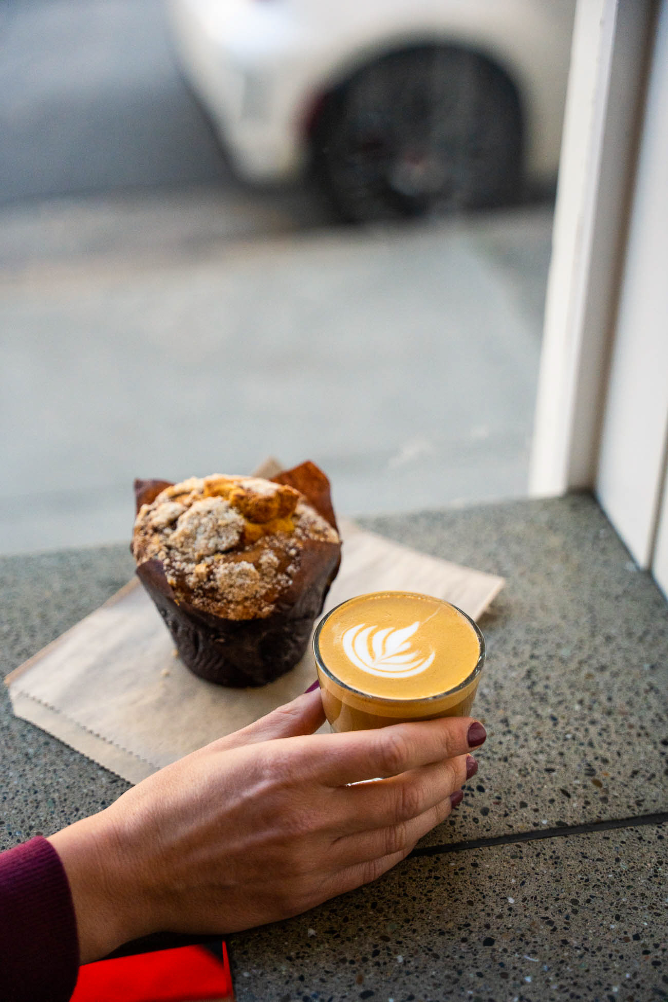 Person's hand holding a cup of coffee with latte art, next to a muffin on a napkin on a windowsill.