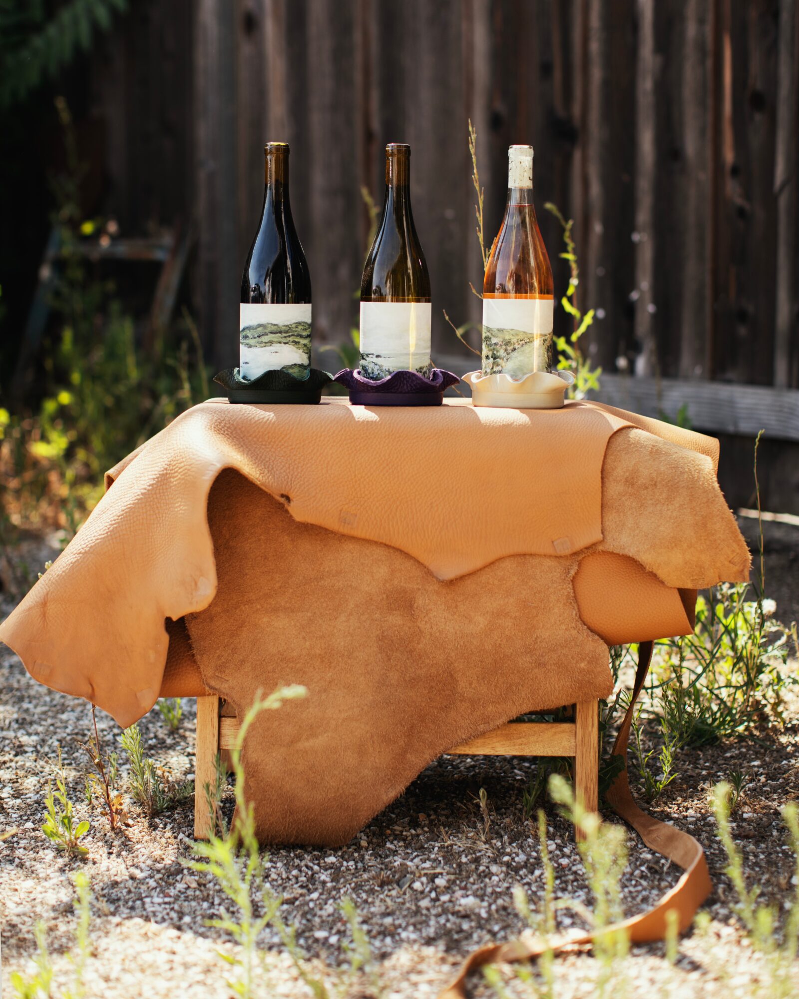 Three bottles of wine on a table covered with a beige cloth, outdoors with a wooden fence in the background.