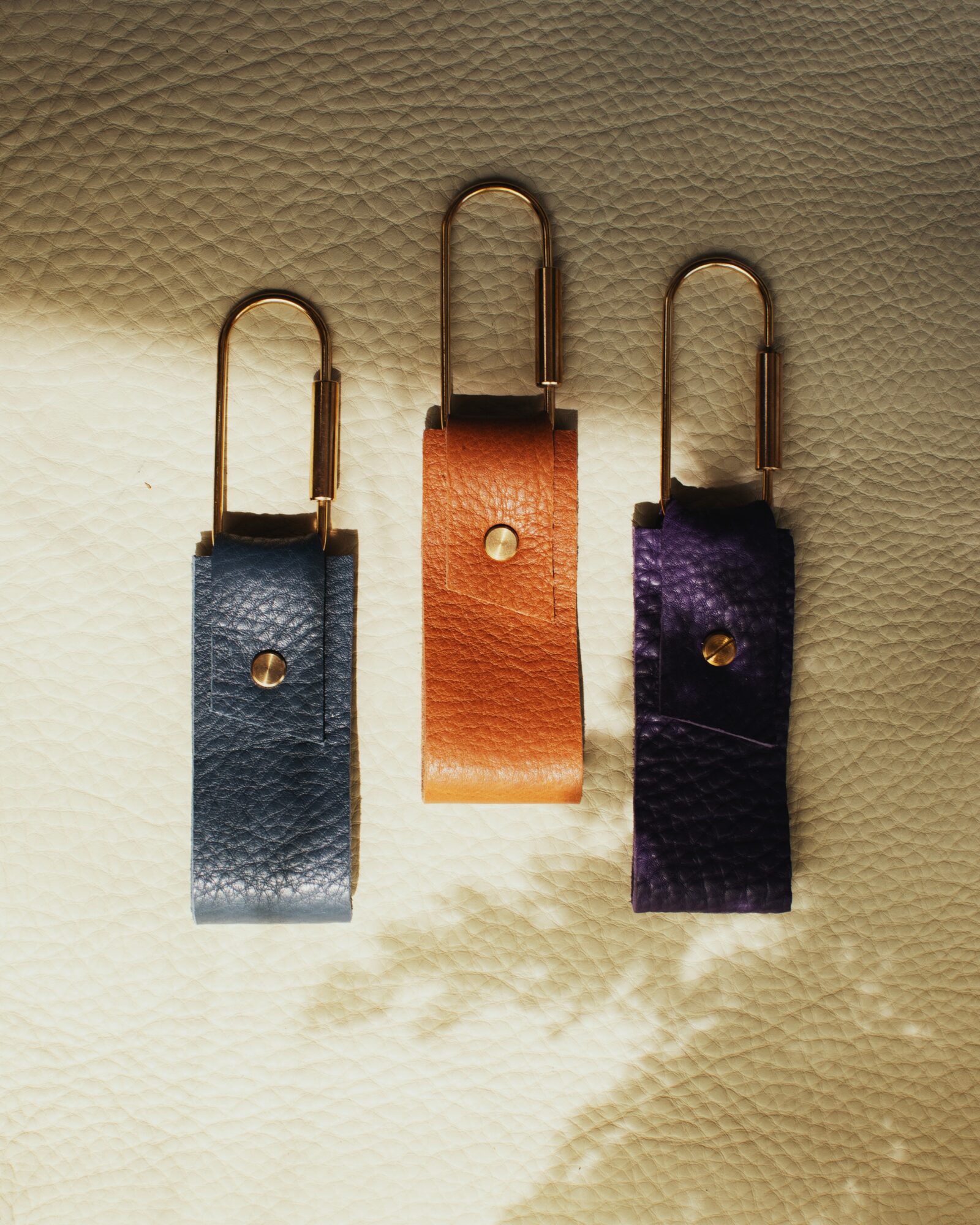 Three leather keychains in blue, orange, and purple with metal hooks, casting shadows on a textured surface.