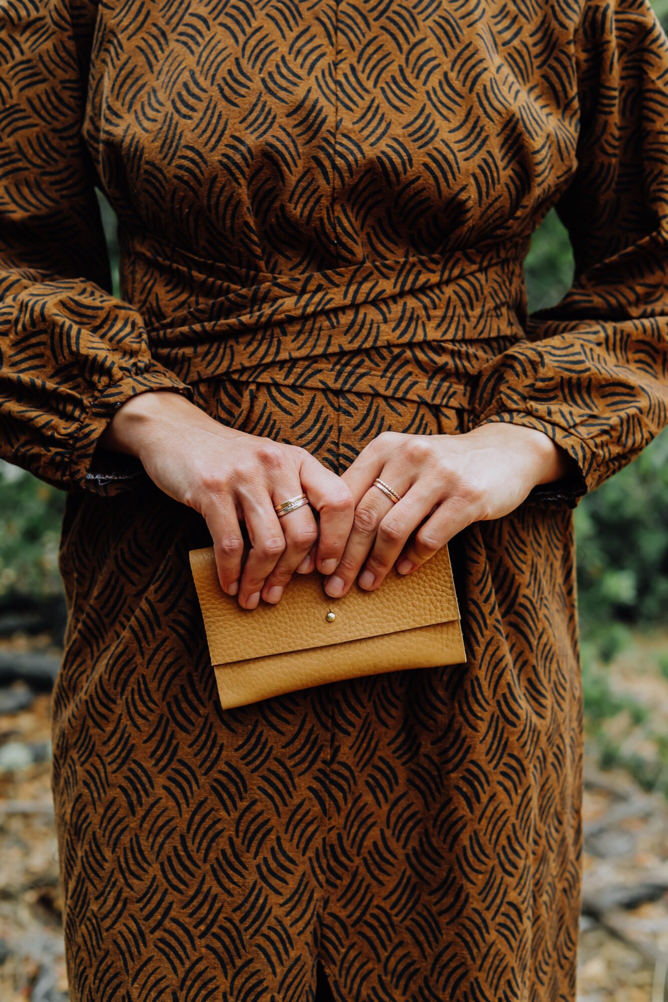 Person in patterned dress holding a small tan clutch with both hands, outdoors background.