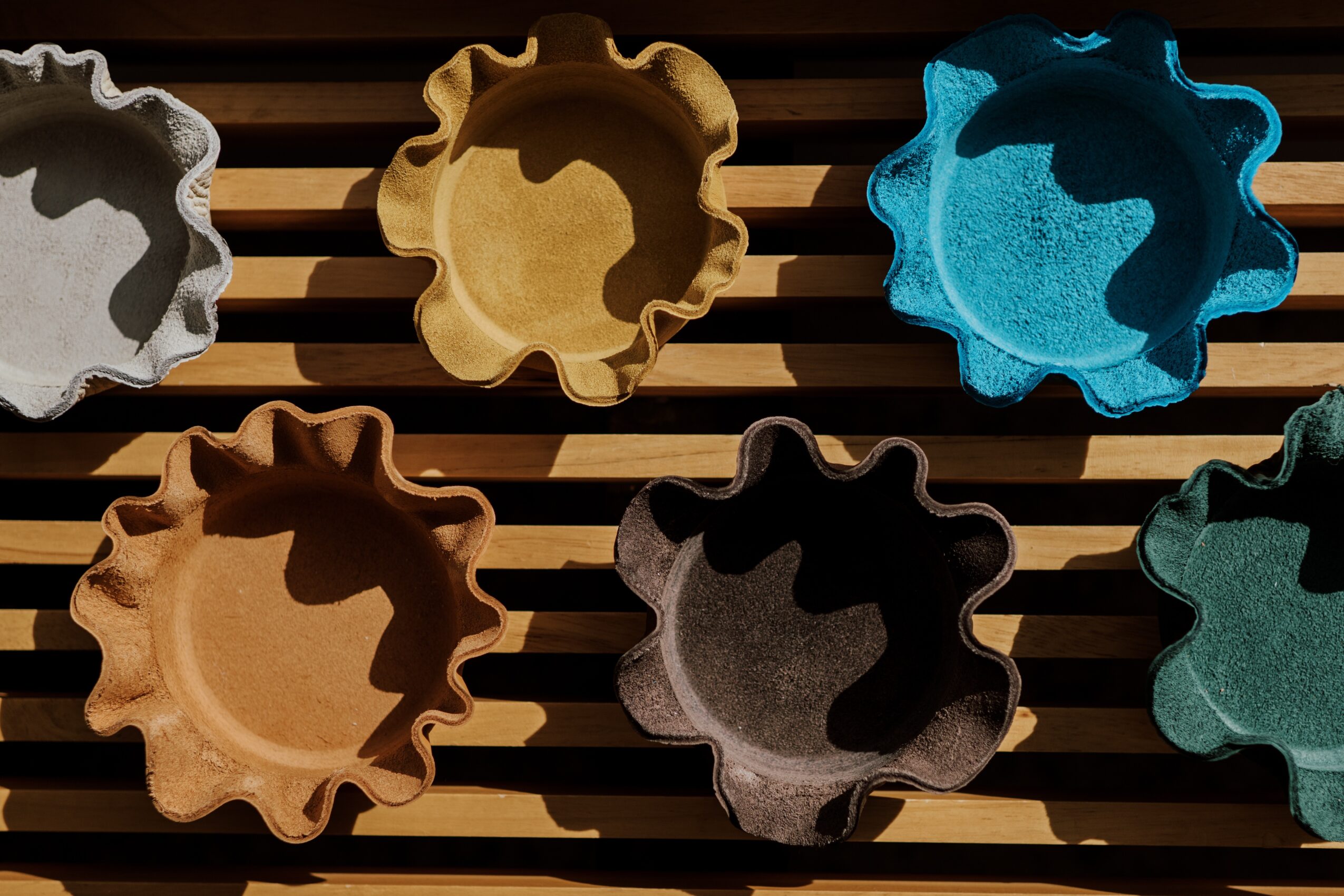 Six colorful ceramic tartlet molds arranged on a wooden slatted surface, with shadows cast by sunlight.