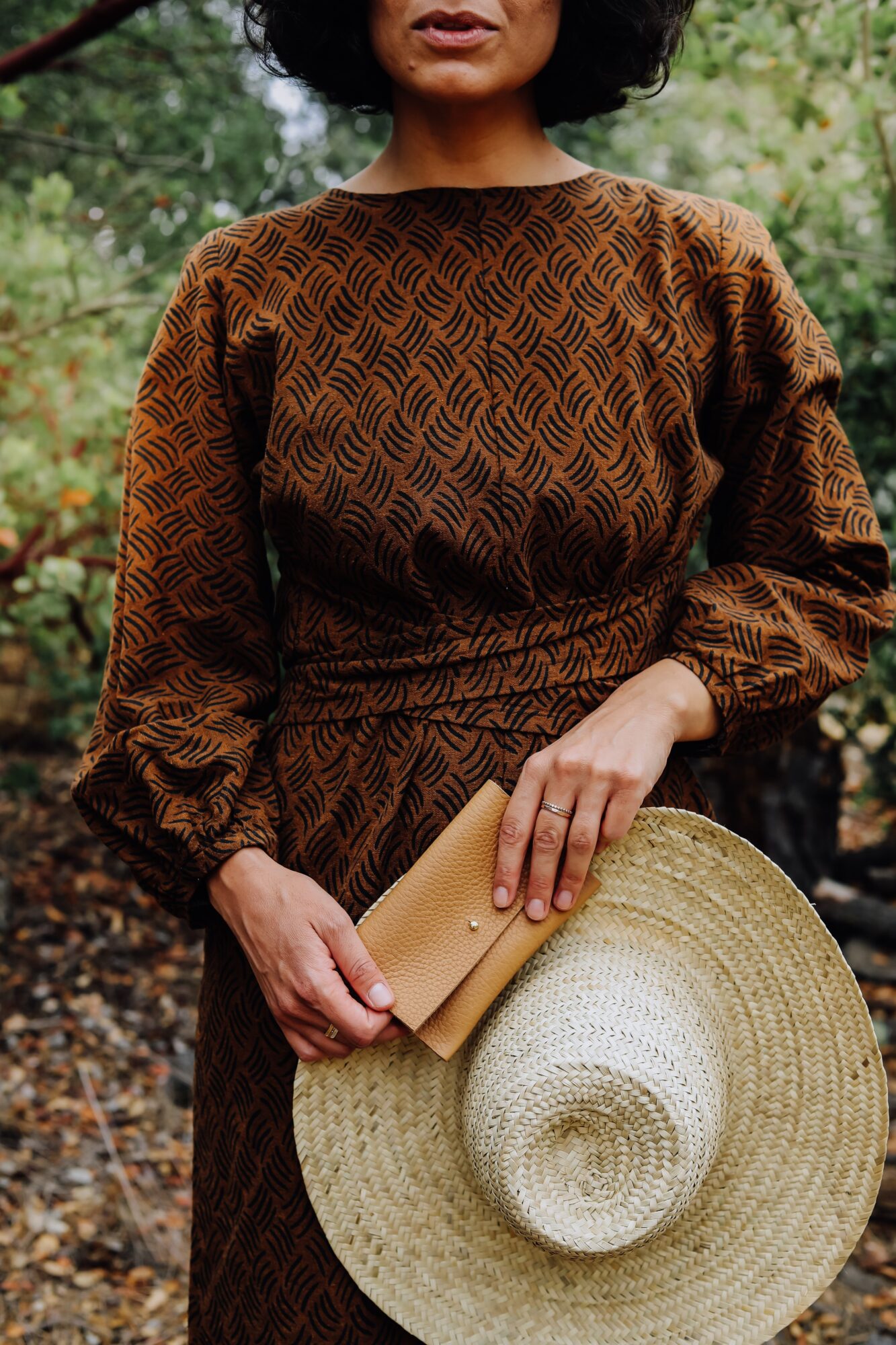 Woman holding a large straw hat and a beige clutch bag outdoors, wearing a patterned brown dress.