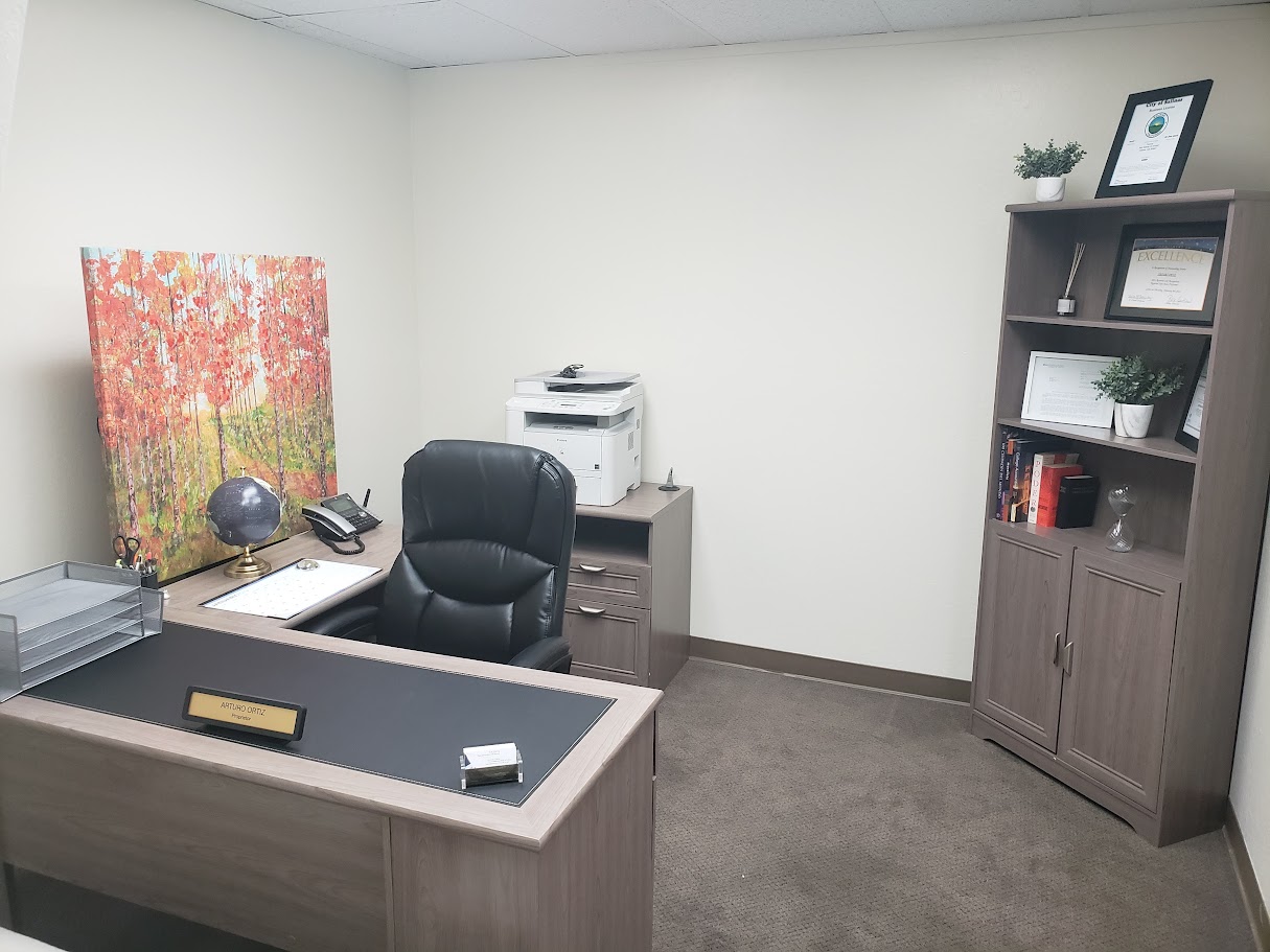 Office room with desk, chair, bookshelf, and printer against plain white walls.