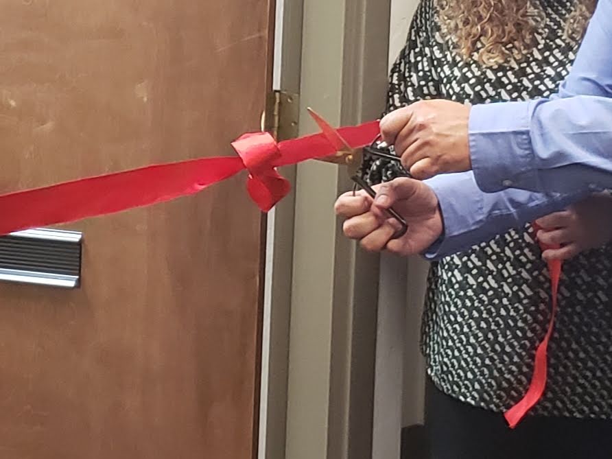 Person cutting a red ribbon with scissors during a ribbon-cutting ceremony, with a woman standing nearby.