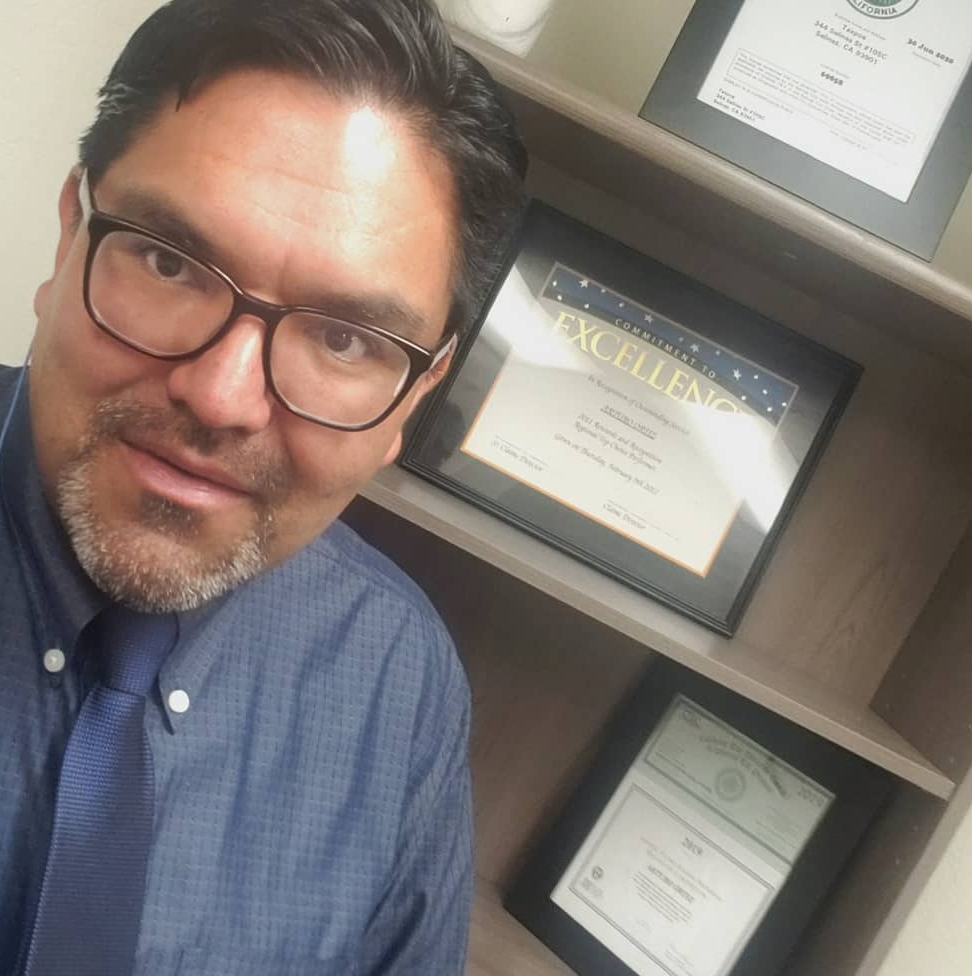 Man with glasses and dark hair takes selfie in front of framed certificates on shelf.