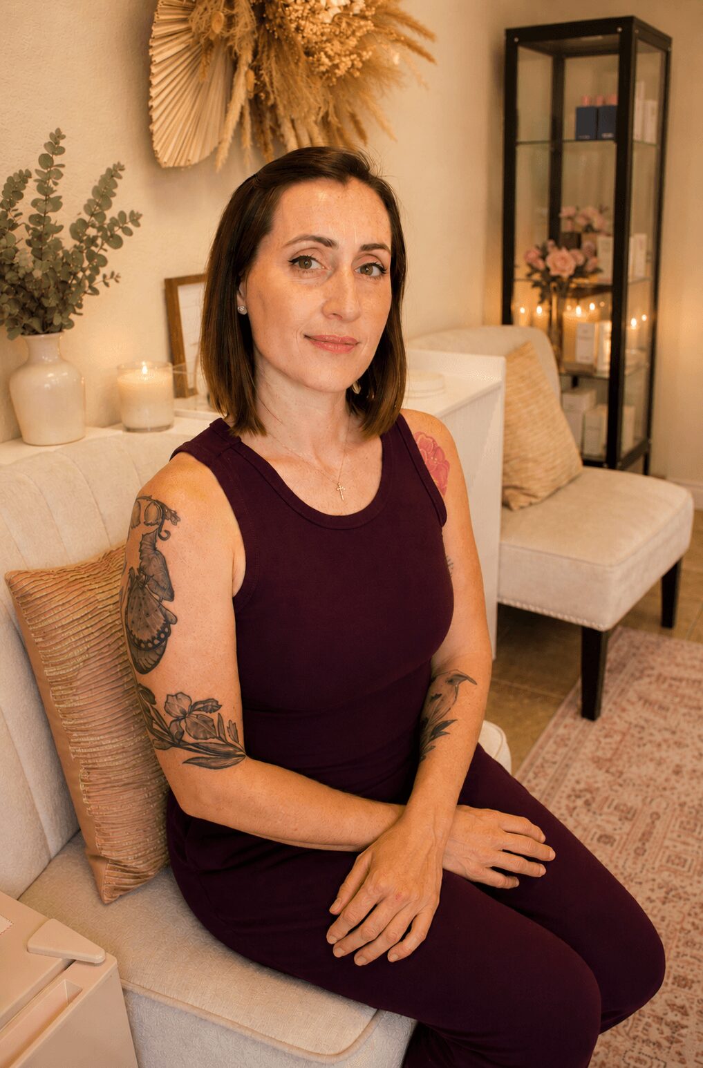 Woman with shoulder-length dark hair and tattoos on arms, sitting on a beige sofa in a cozy room with candles and plants.