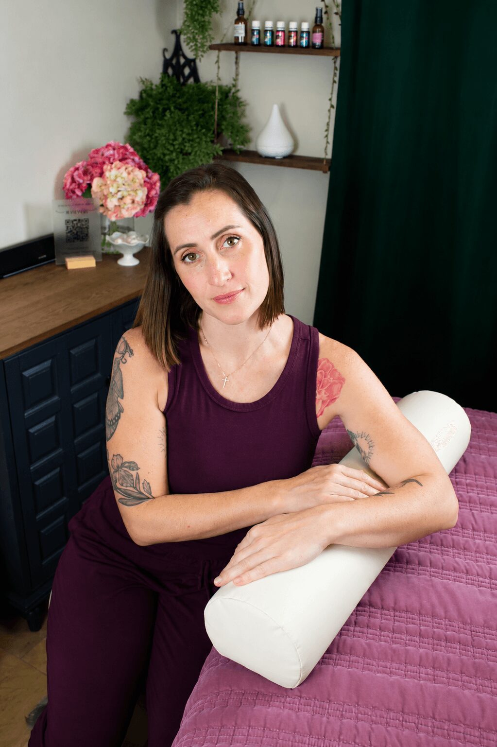 Woman with tattoos leaning on a white pillow on a bed, in a room with flowers and shelves in the background.