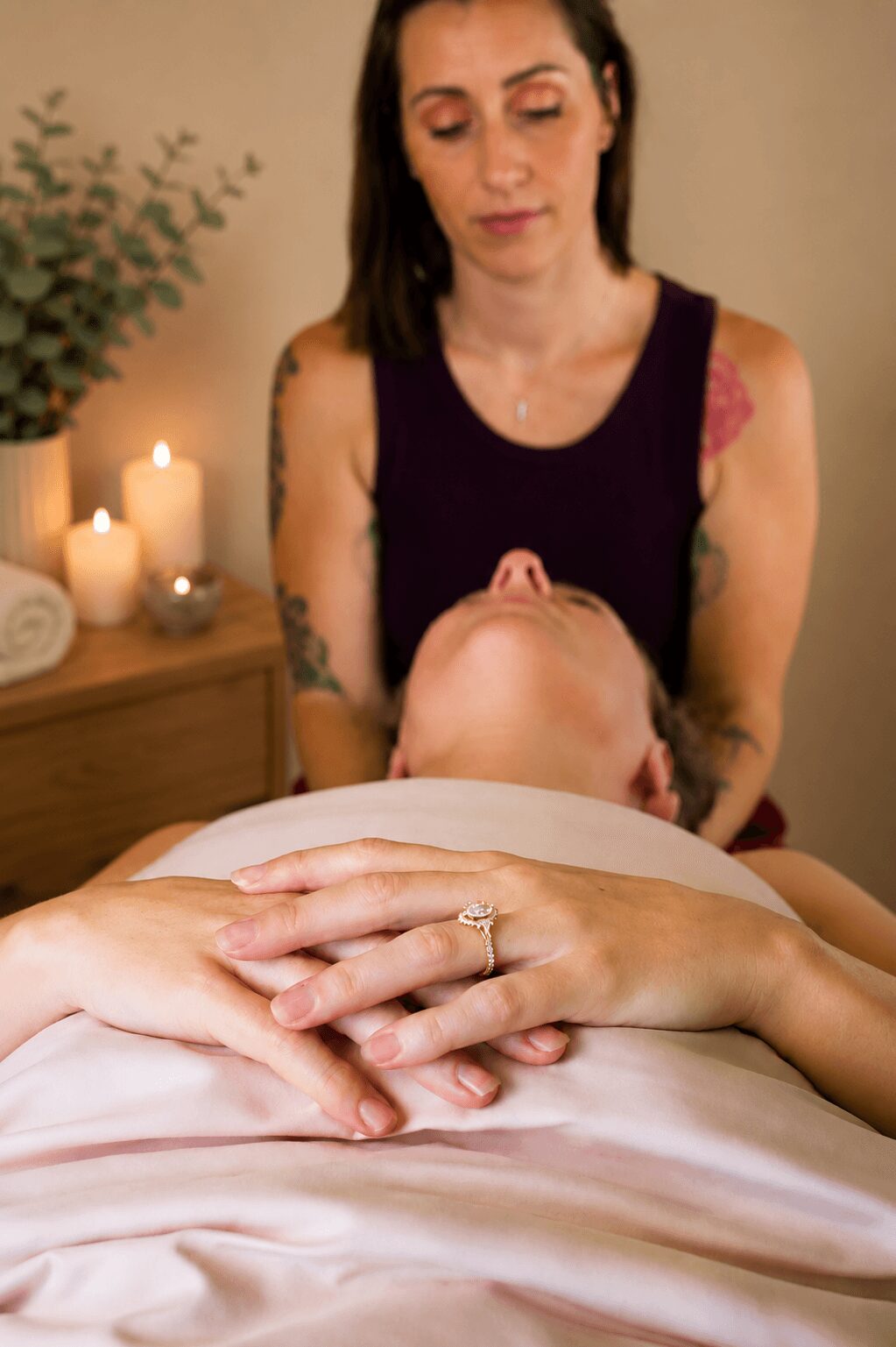 Woman with tattoos and dark hair looks down at a person receiving a massage, hands resting on the bed, candles in background.