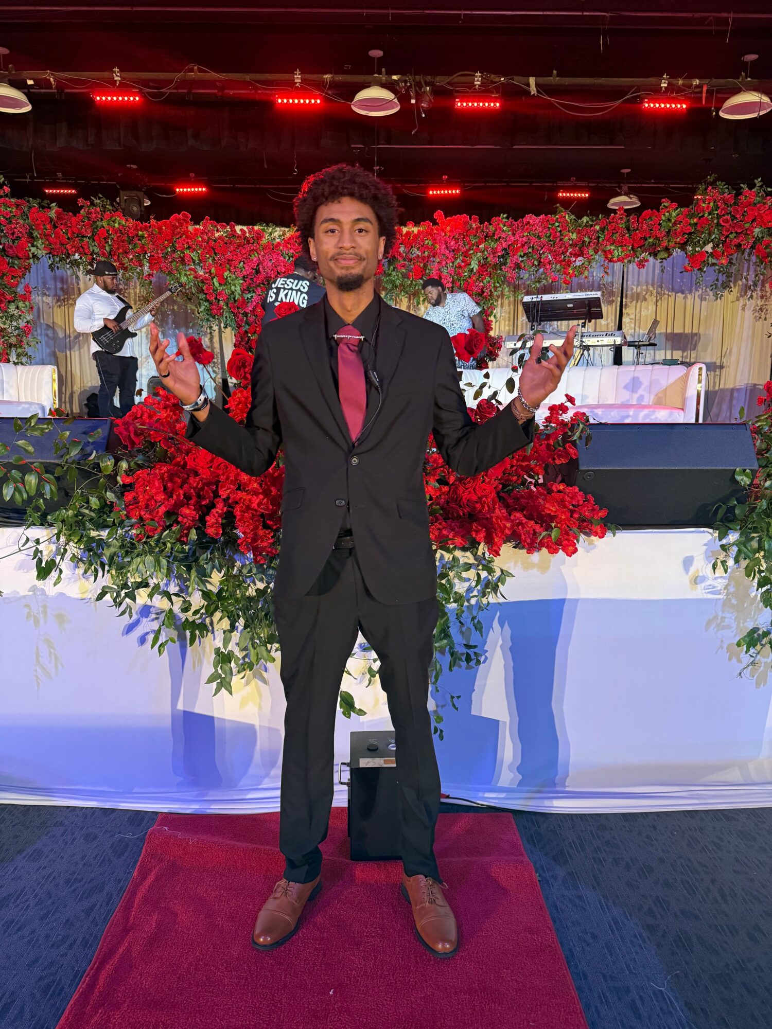 Person in a black suit standing on a red carpet with floral arrangements and a decorated background.
