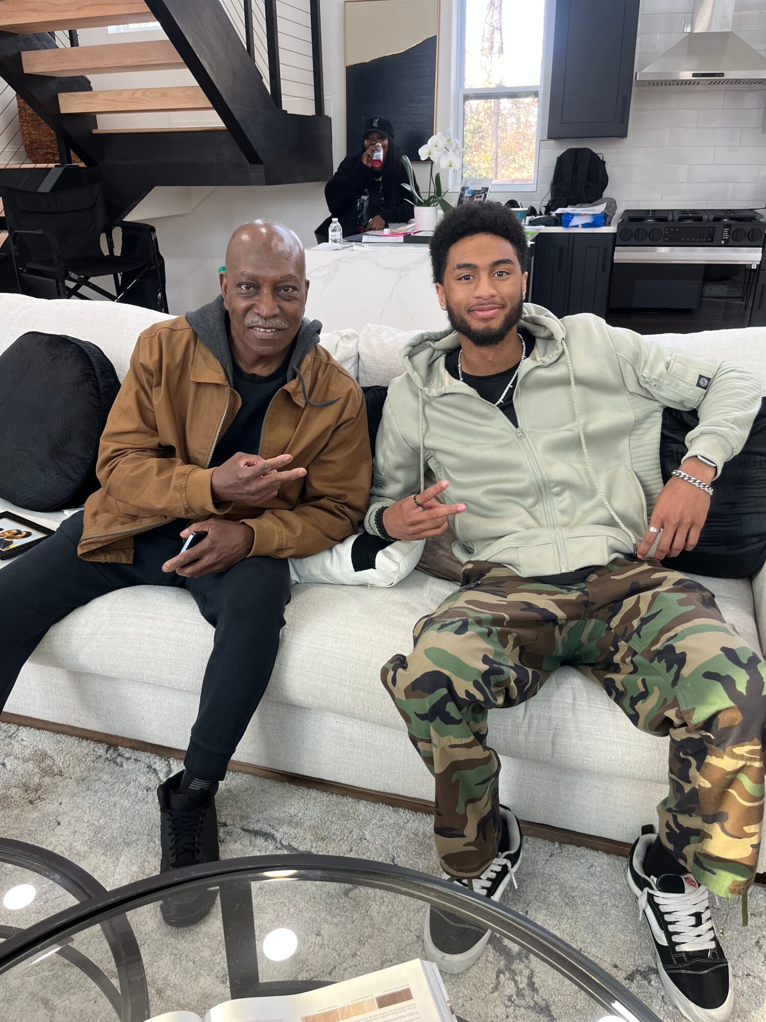 Two men sitting on a white couch, one in a white hoodie and camouflage pants, the other in a brown jacket, both making peace signs.