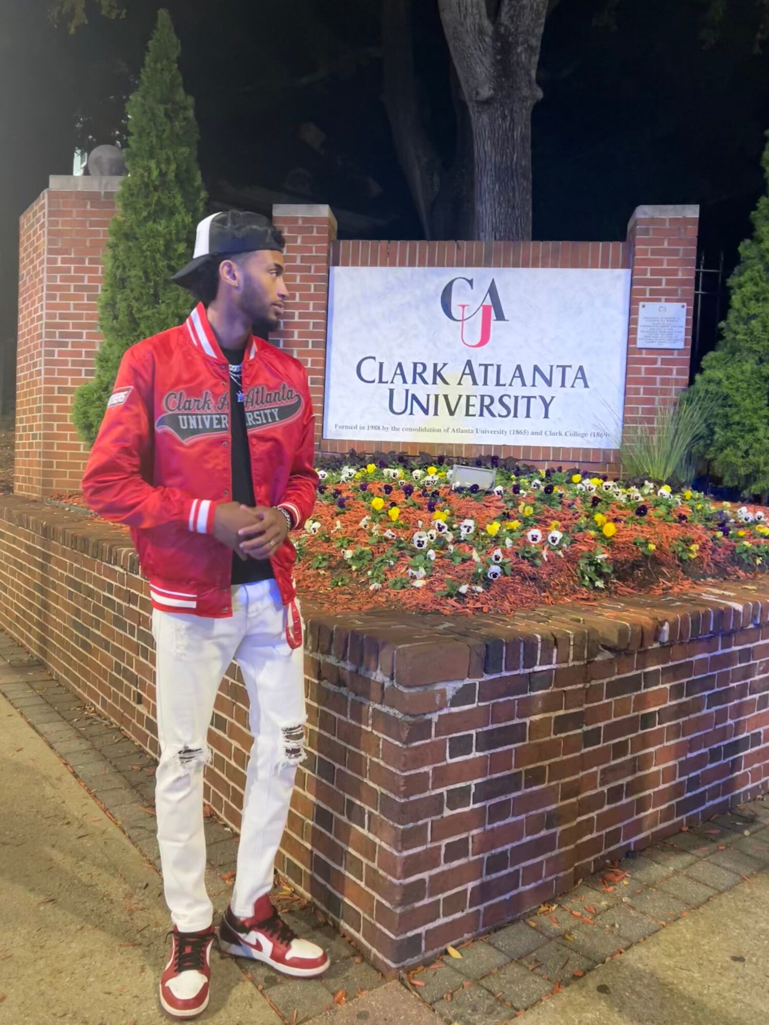 Person wearing a red jacket, white pants, and a hat standing near a brick wall and Clark Atlanta University sign at night.
