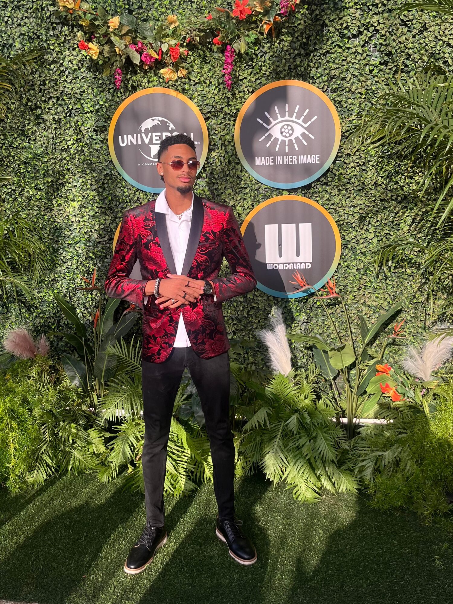 Person in red blazer and sunglasses standing outdoors with green plants and three circular signs behind them.