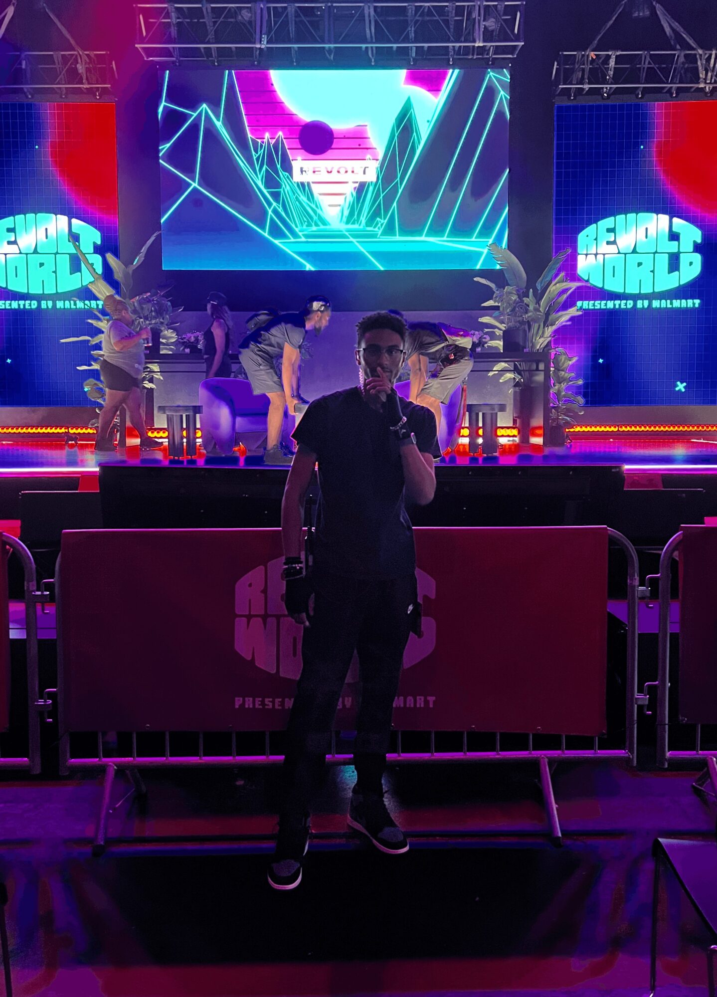 Person standing in front of a stage with colorful digital screens and performers, illuminated by purple and blue lights.