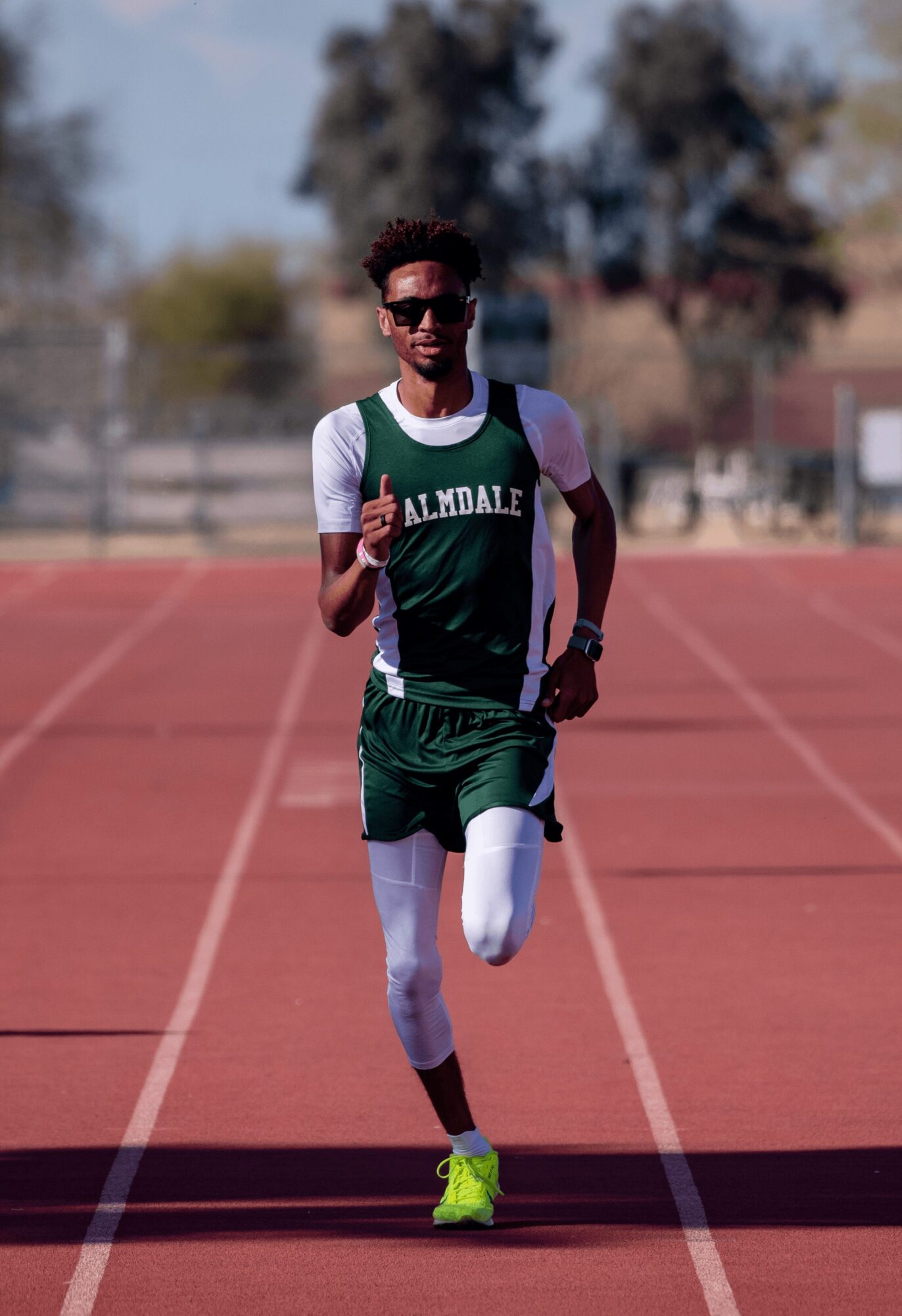 Young male athlete running on a track, wearing a green and white uniform with yellow shoes, outdoors on a sunny day.