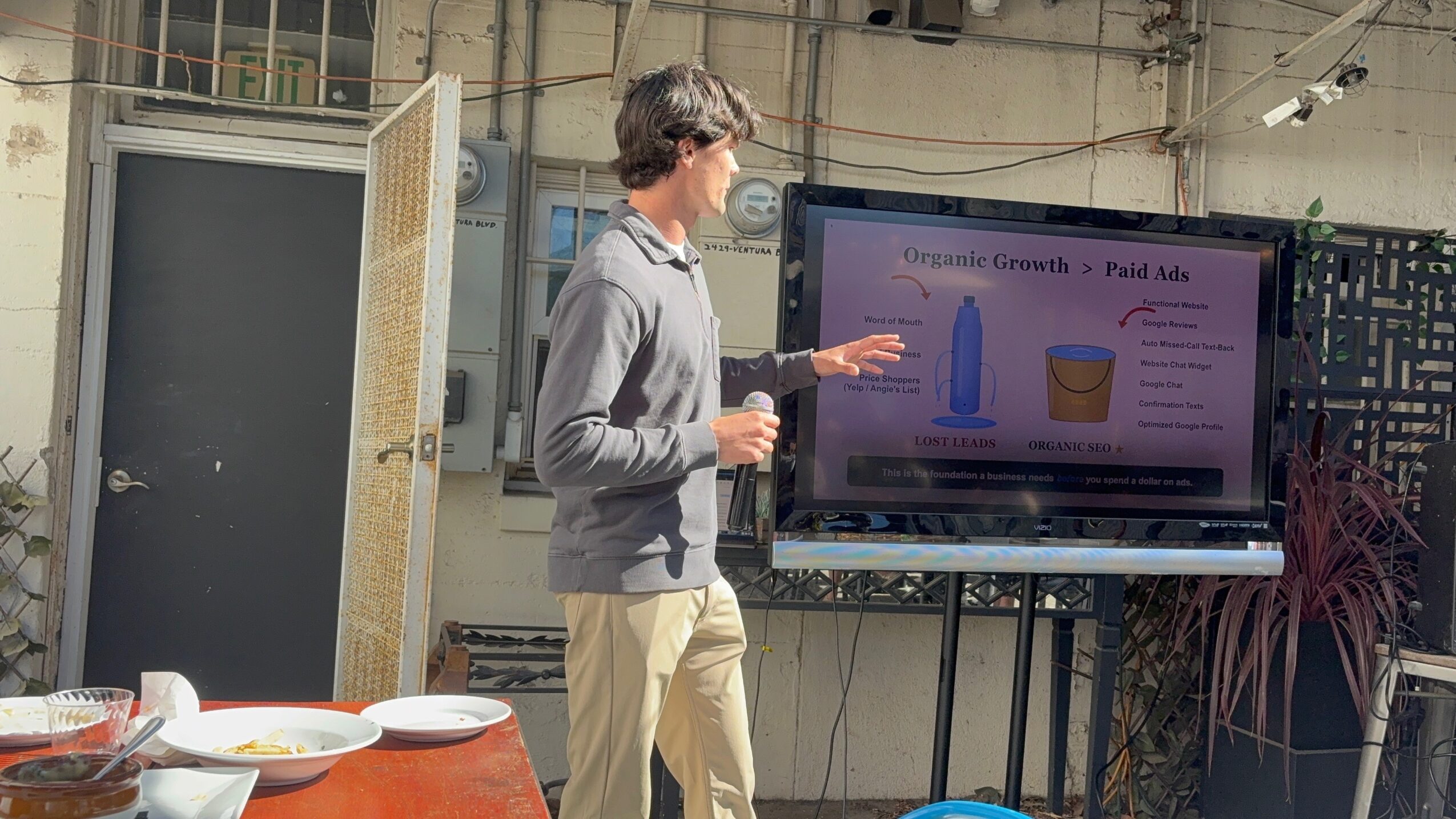 Person presenting in front of a large screen with a graph and text, in an industrial-style room with plants and tables.