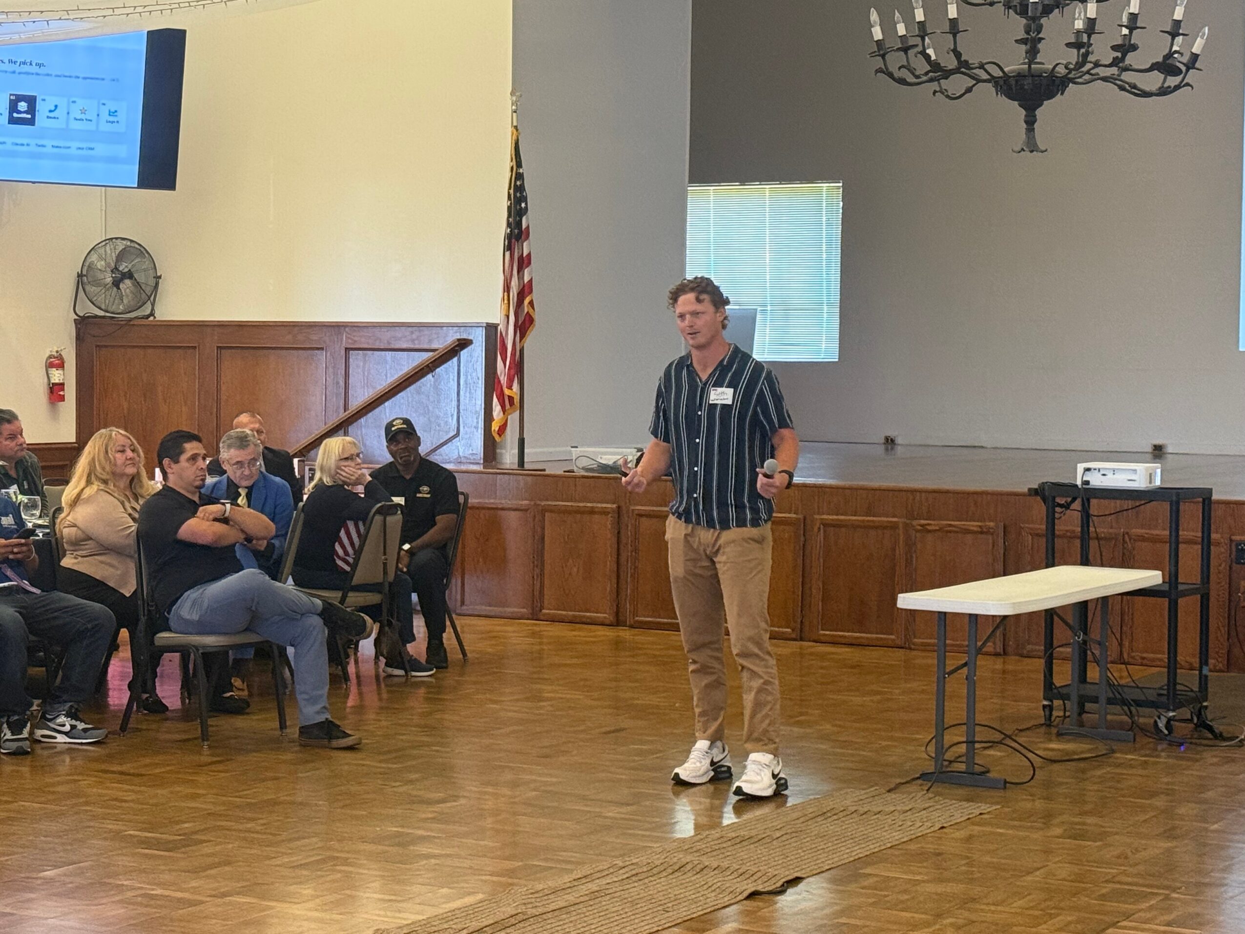 Young man standing and speaking to a group in a room with wooden floors and a chandelier.