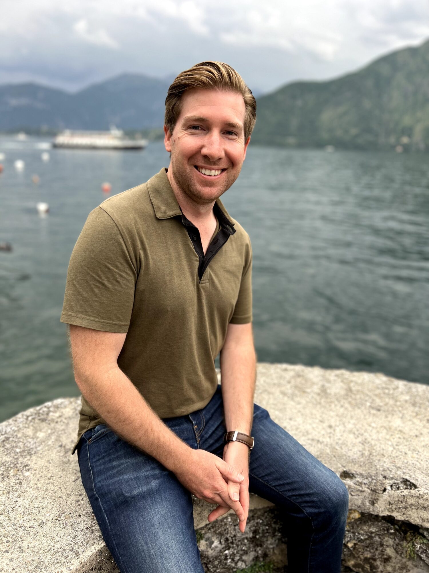 Smiling man sitting on a rock near water with mountains in the background.
