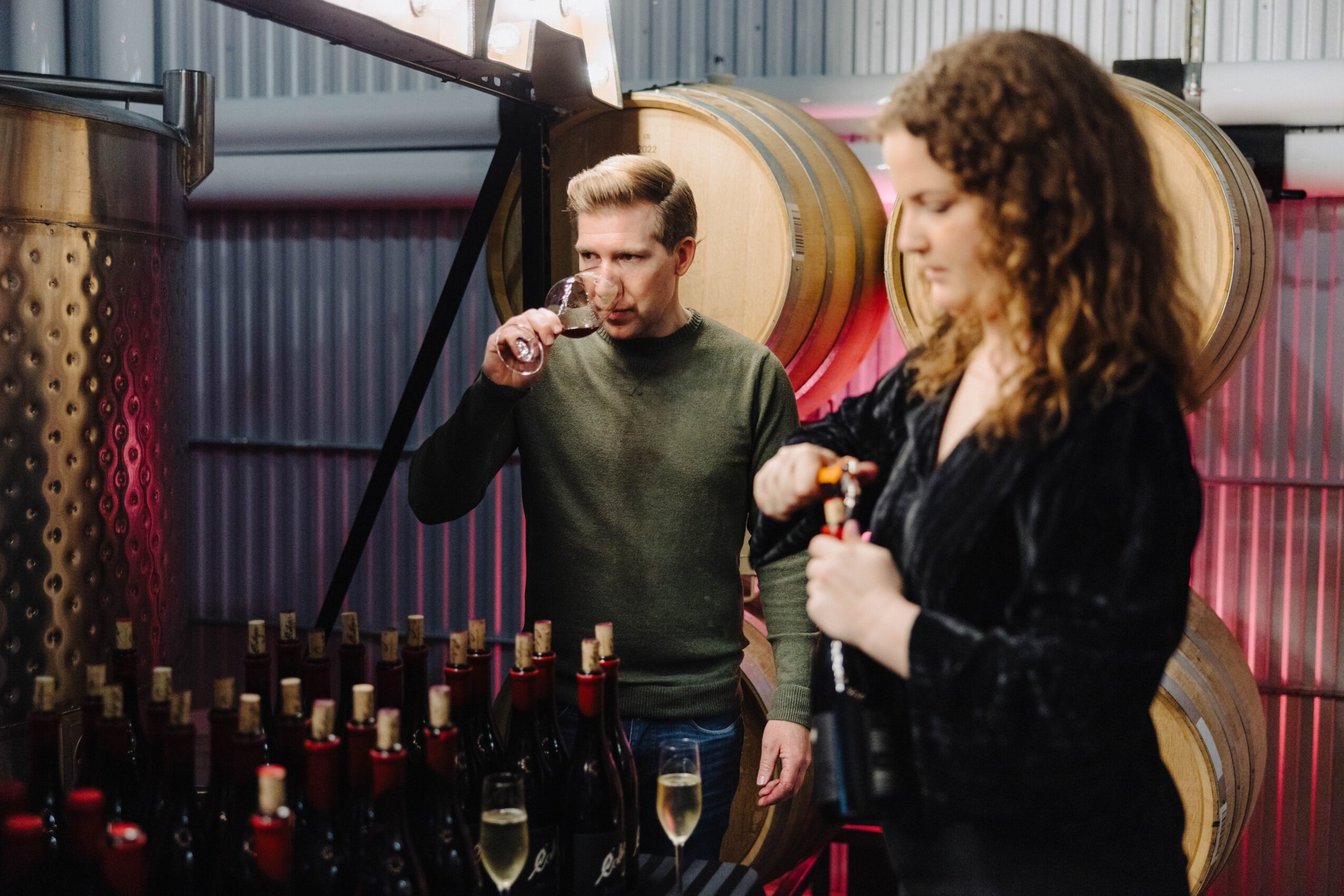 A man and woman tasting wine in a winery with wine bottles and barrels in the background.