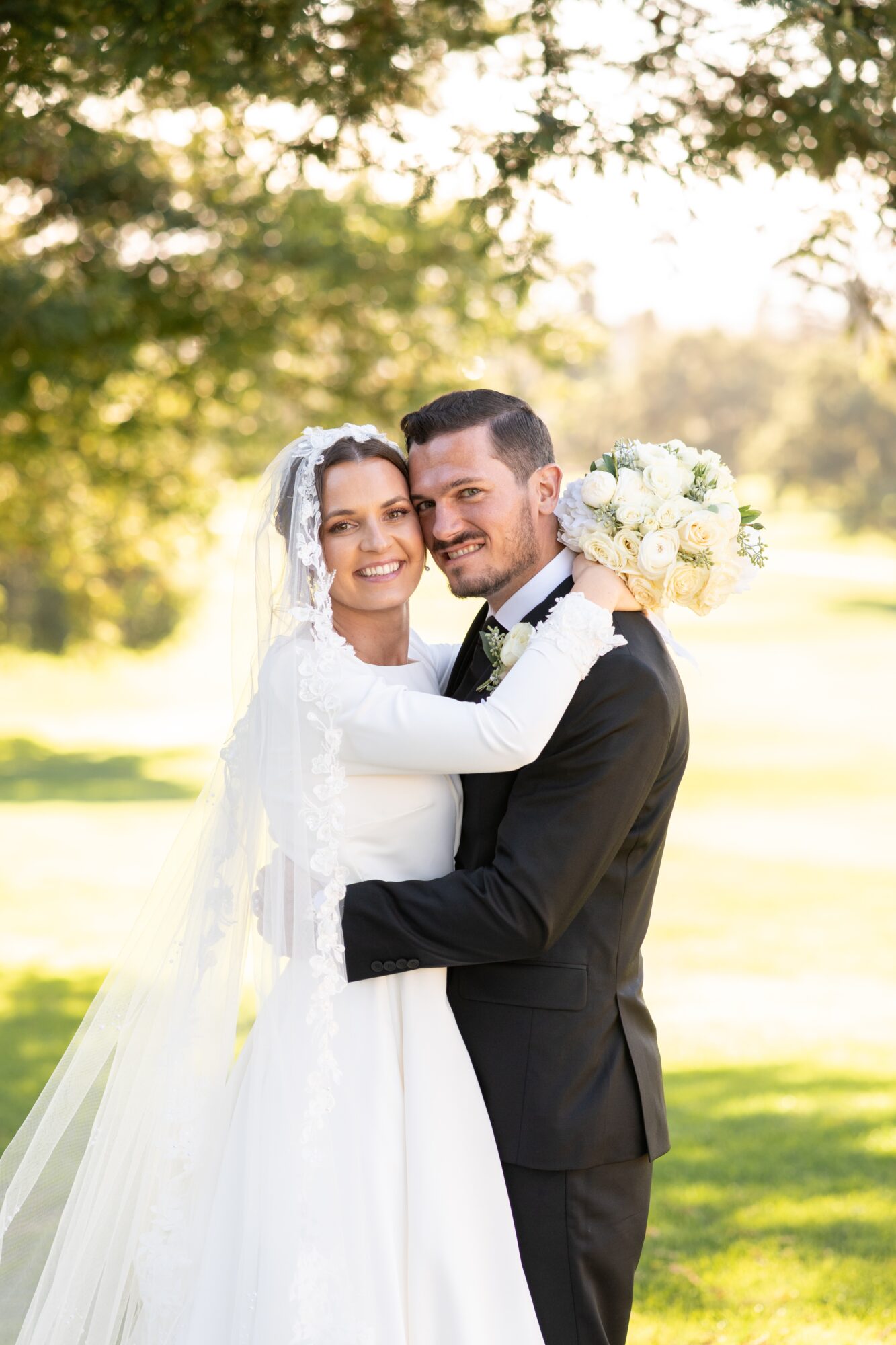 Bride and groom smiling outdoors, bride holding a bouquet, trees and sunlight in background