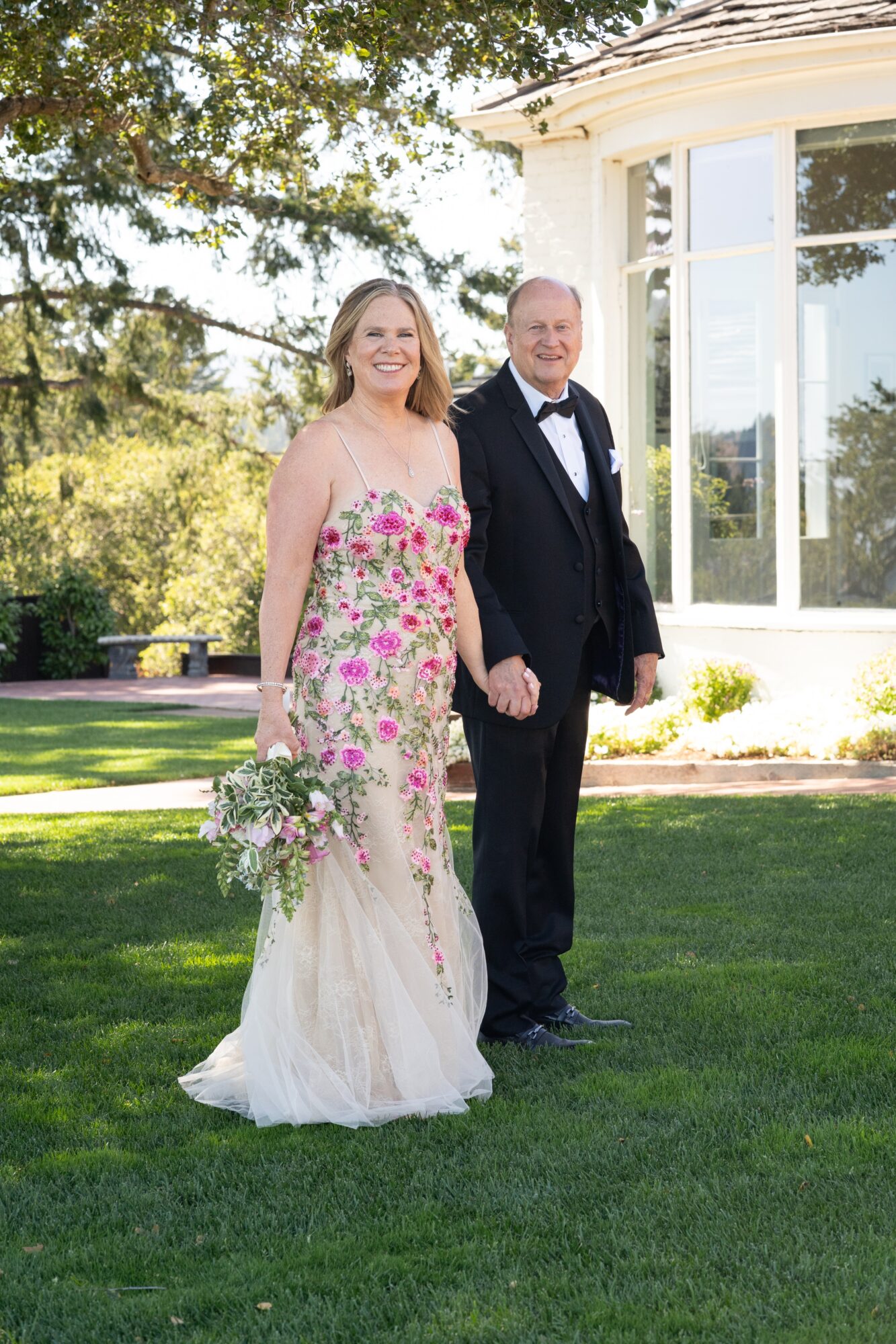 A bride and groom stand outdoors on grass, smiling, with trees and a white building in the background.