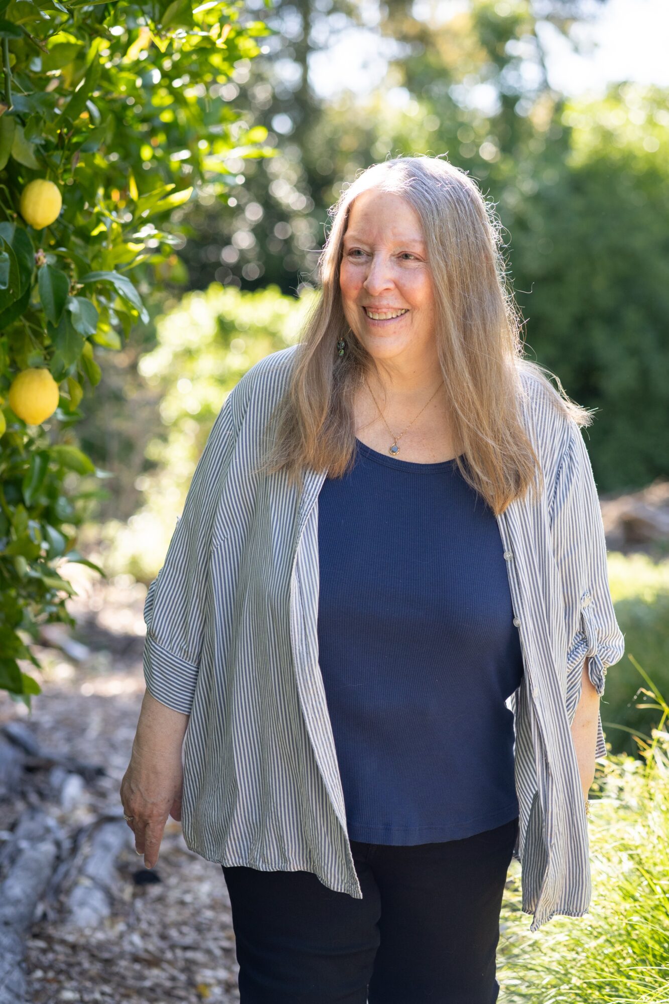 Smiling woman with long hair outdoors near lemon trees, sunlight filtering through leaves, wearing a blue top and light jacket.