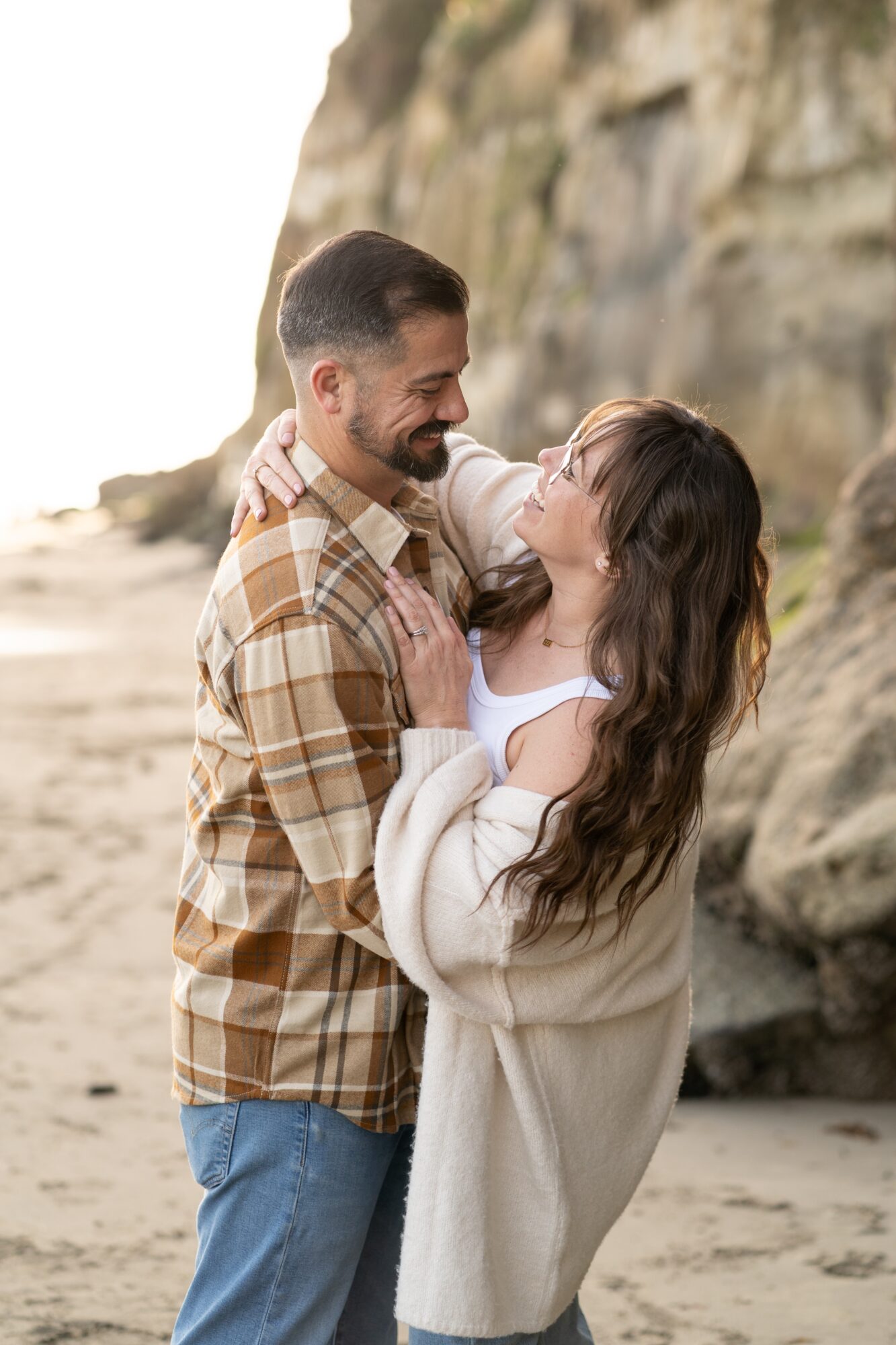 A smiling couple stands close together on a beach, with rocky cliffs in the background, embracing each other.
