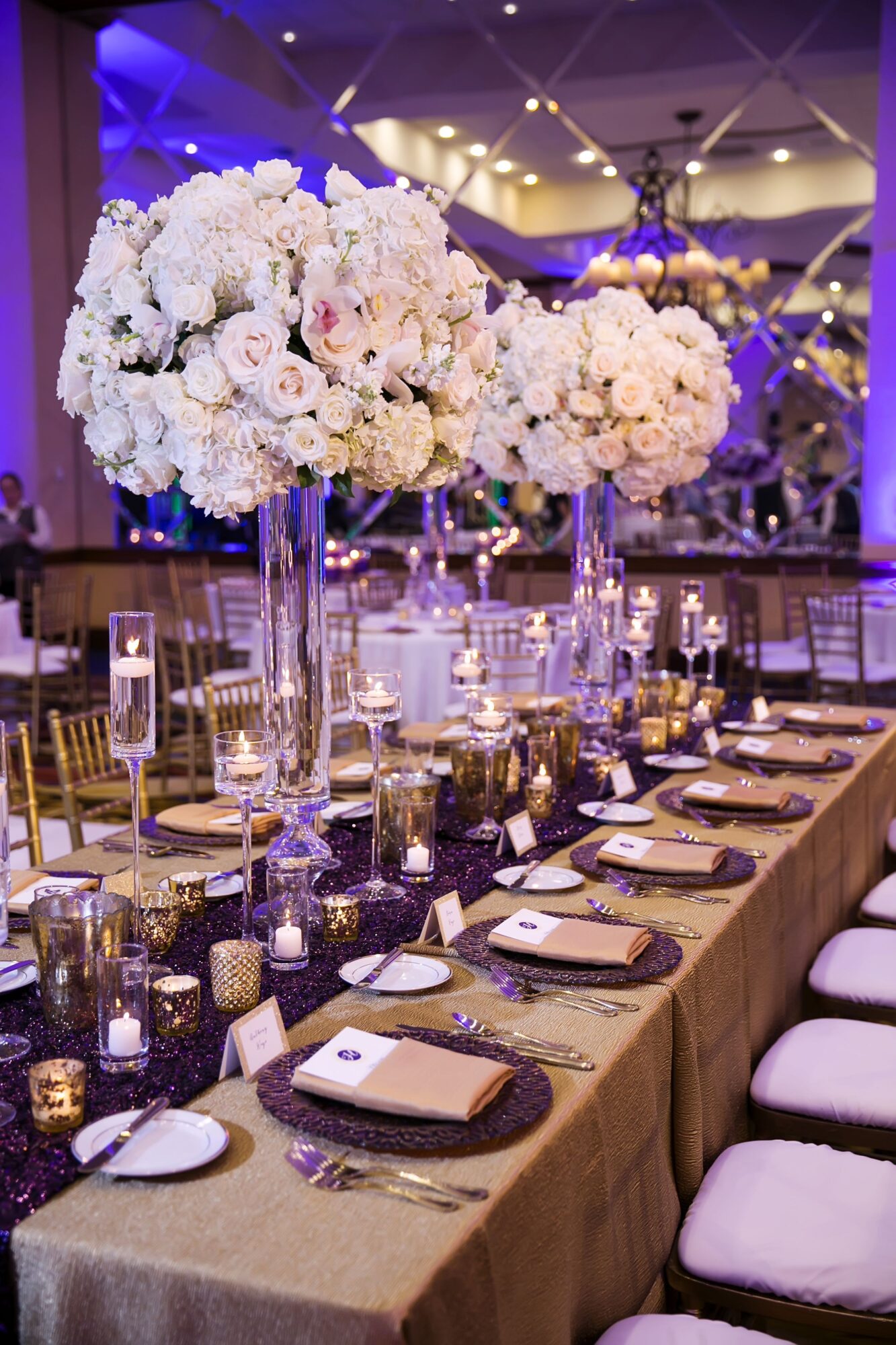 Long banquet table with tall floral centerpieces of white flowers, set with plates, glasses, and candles in a decorated event space.