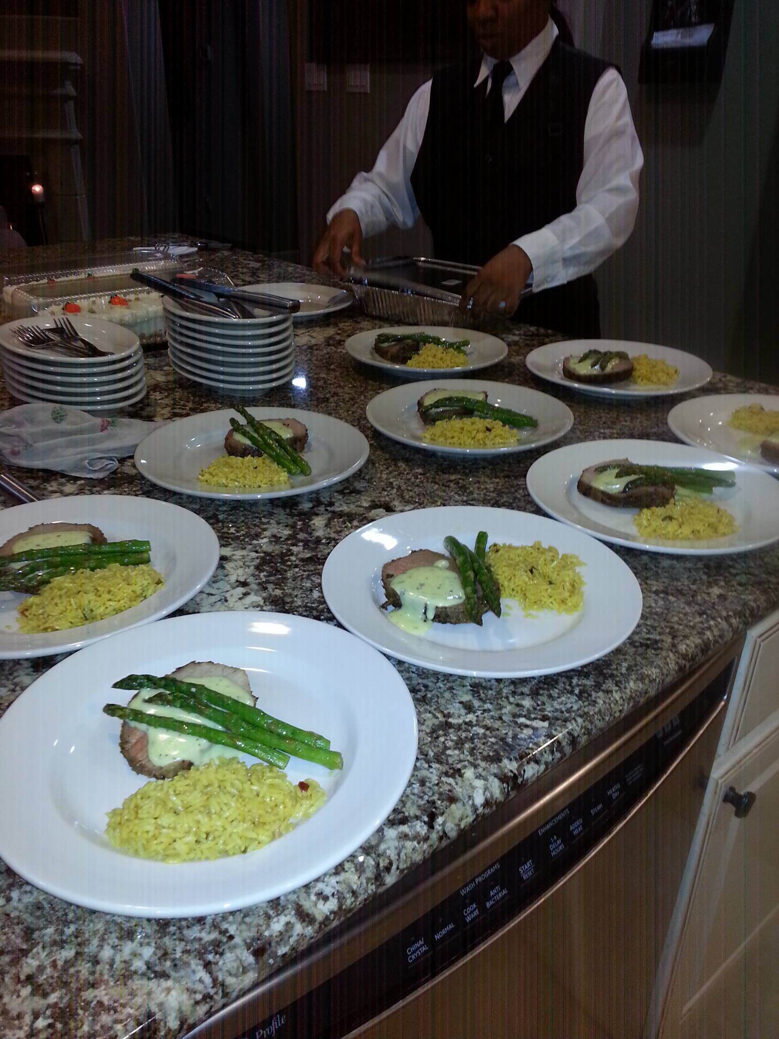 Multiple plates with rice, green vegetables, and fish on a granite countertop, with a person in formal attire in the background.