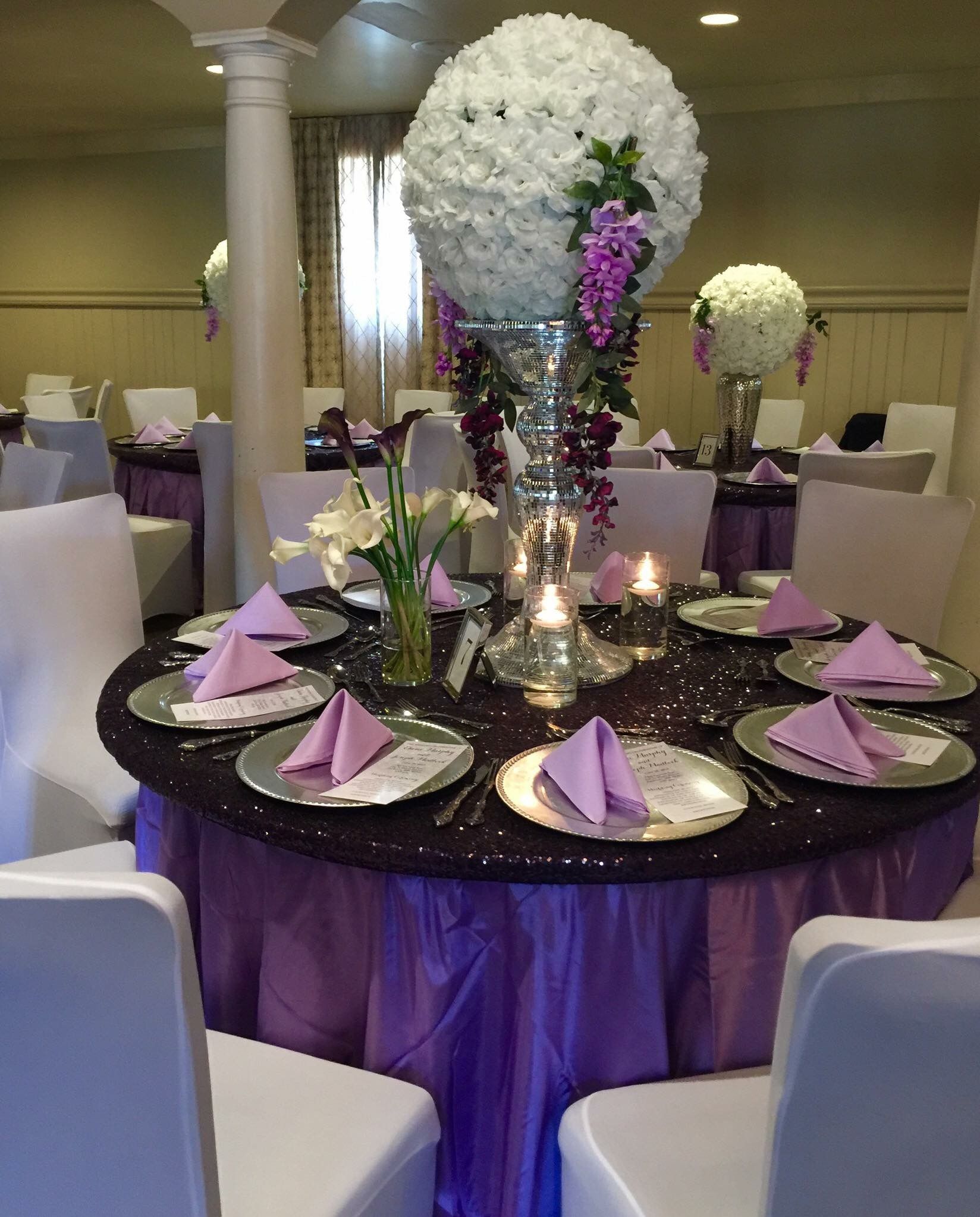 Round table with purple tablecloth, white chairs, floral centerpiece, candles, and purple napkins, set for a formal event.