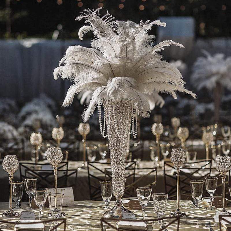 Tall centerpiece with white feathers and beads on a decorated table at an event, with chairs and tableware visible.