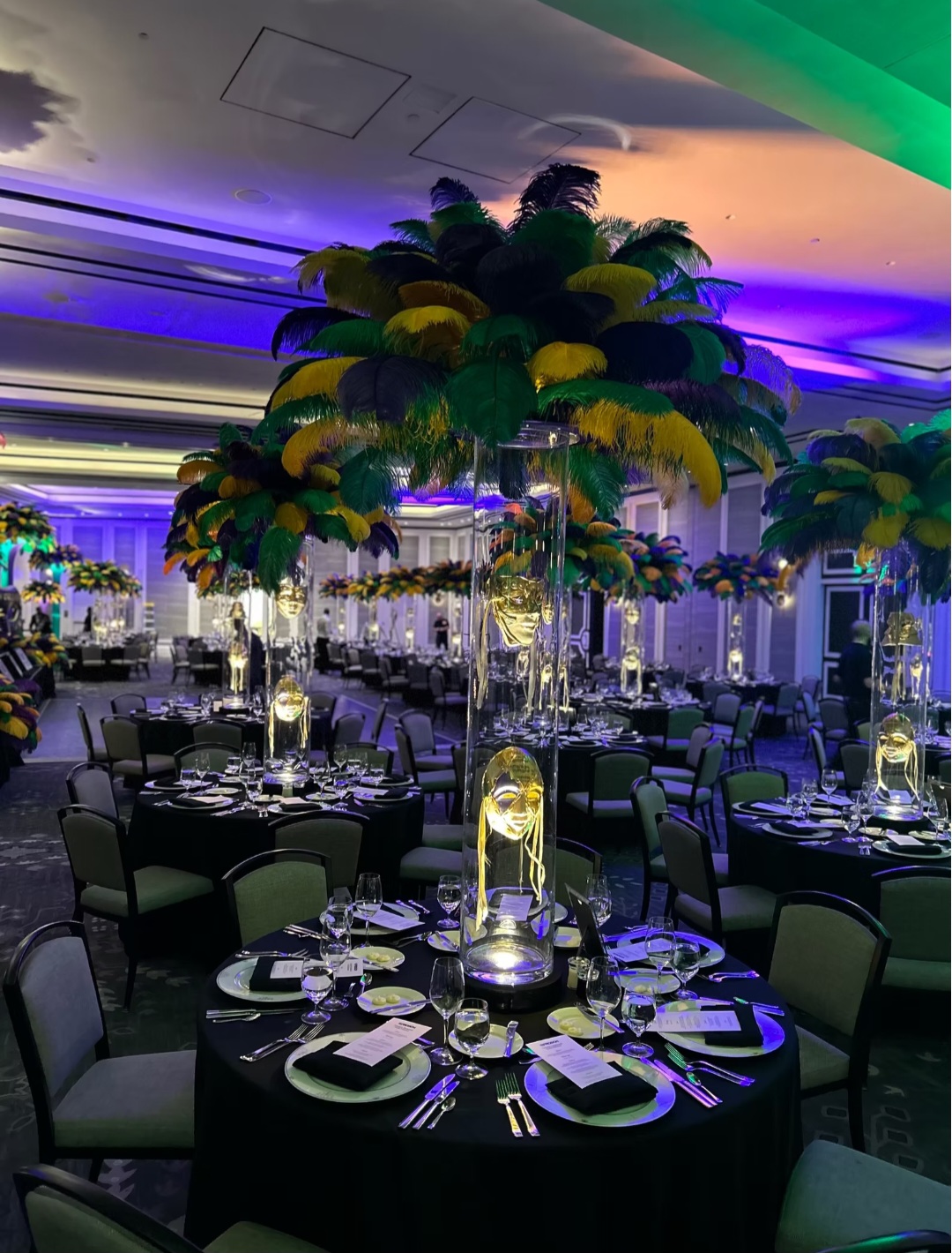 Elegant event space with round tables, black tablecloths, tall floral centerpieces, and ambient lighting in purple and green hues.